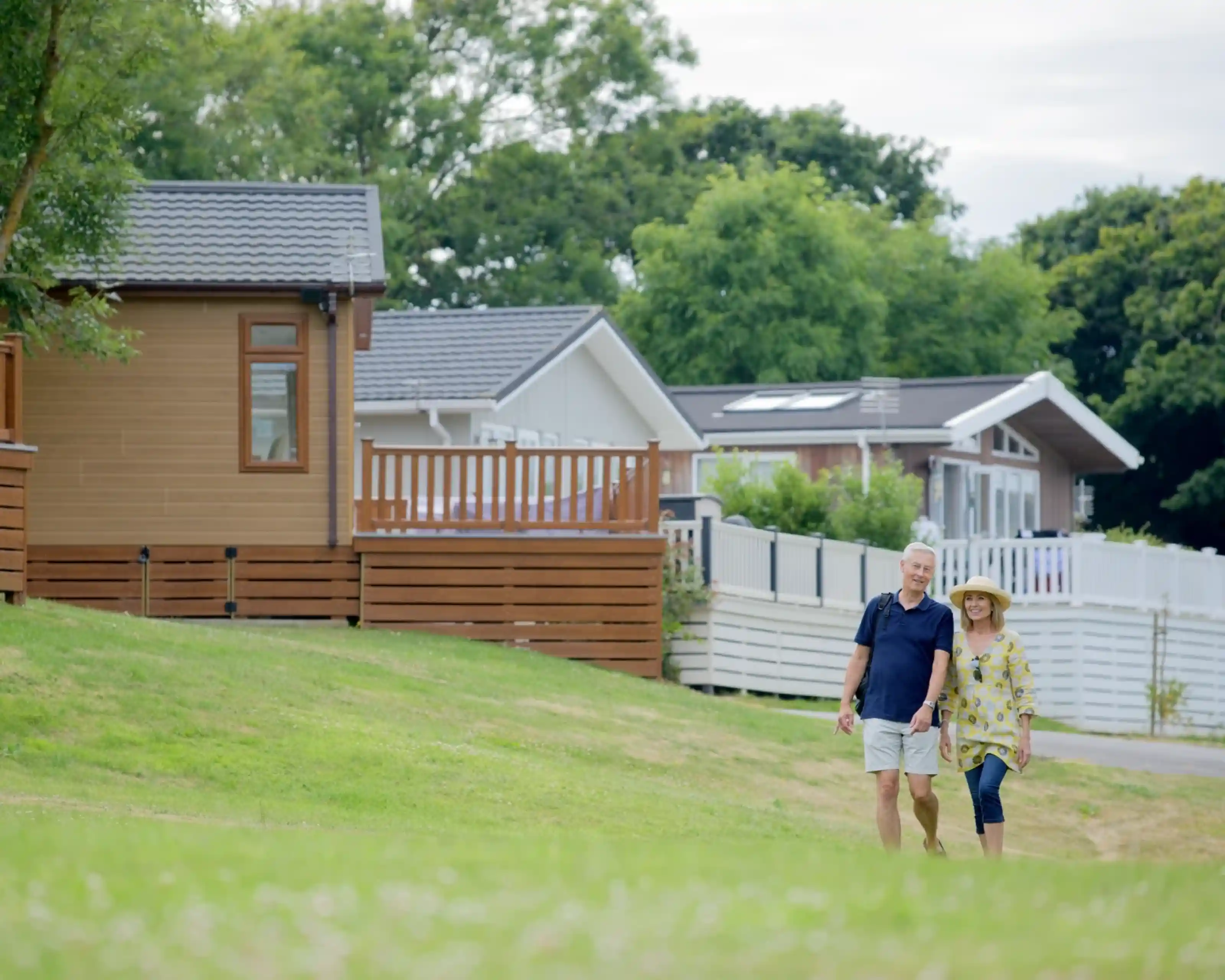 An elderly couple walks hand in hand along a grassy path, with modern, wooden vacation homes in the background surrounded by lush greenery.