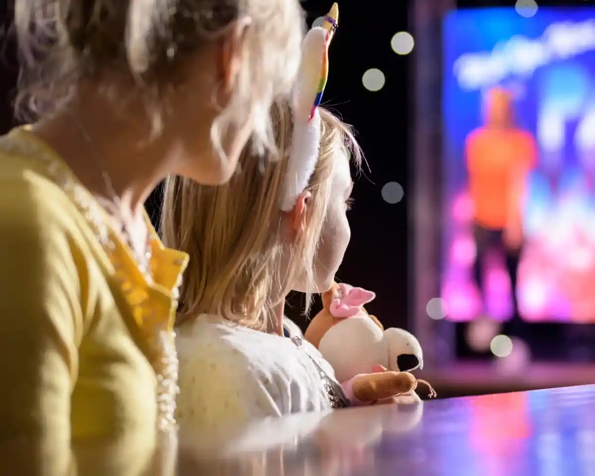 Two children watch a performance from a distance, one wearing a unicorn headband and the other holding a stuffed animal. The stage is illuminated with colorful lights and a performer can be seen in the background.