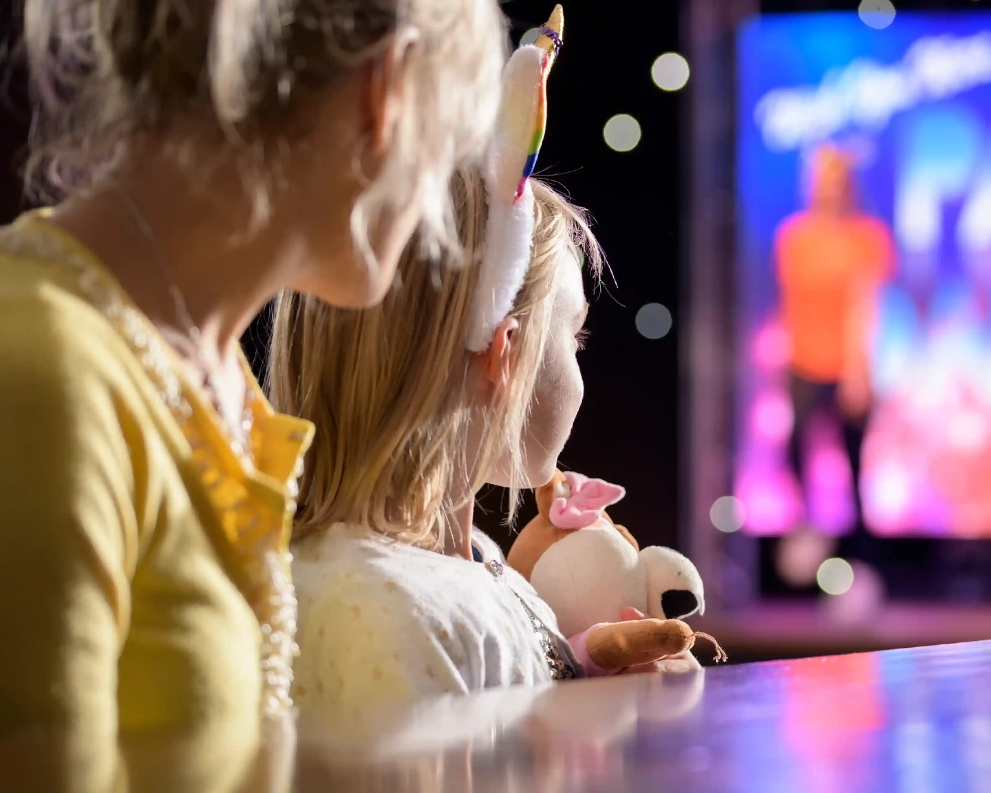 Two children watch a performance from a distance, one wearing a unicorn headband and the other holding a stuffed animal. The stage is illuminated with colorful lights and a performer can be seen in the background.