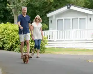 A couple walks together on a path, holding hands and smiling. A small dog on a leash leads the way in front of them. In the background, a cozy white cabin is visible, surrounded by greenery.