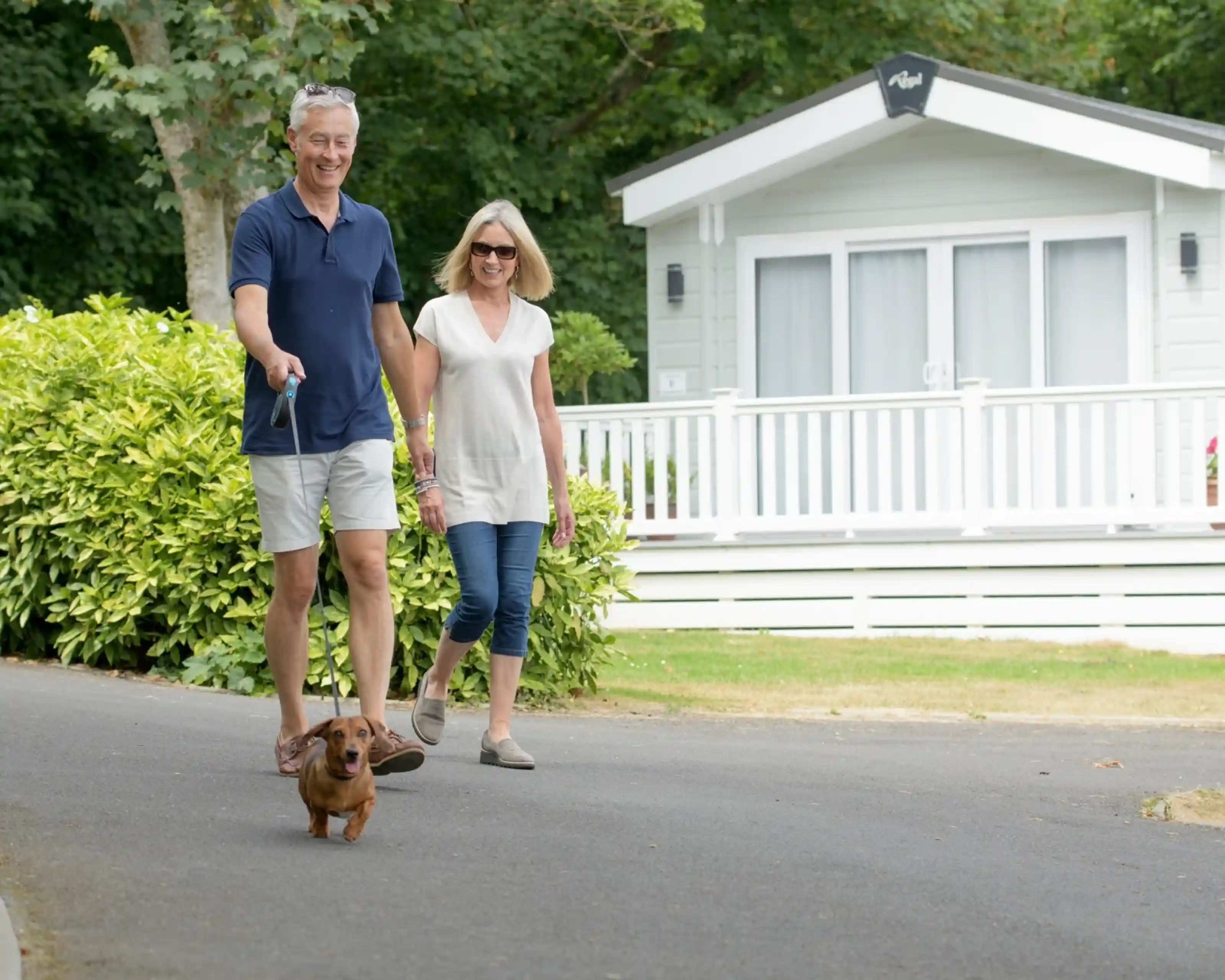 A couple walks together on a path, holding hands and smiling. A small dog on a leash leads the way in front of them. In the background, a cozy white cabin is visible, surrounded by greenery.