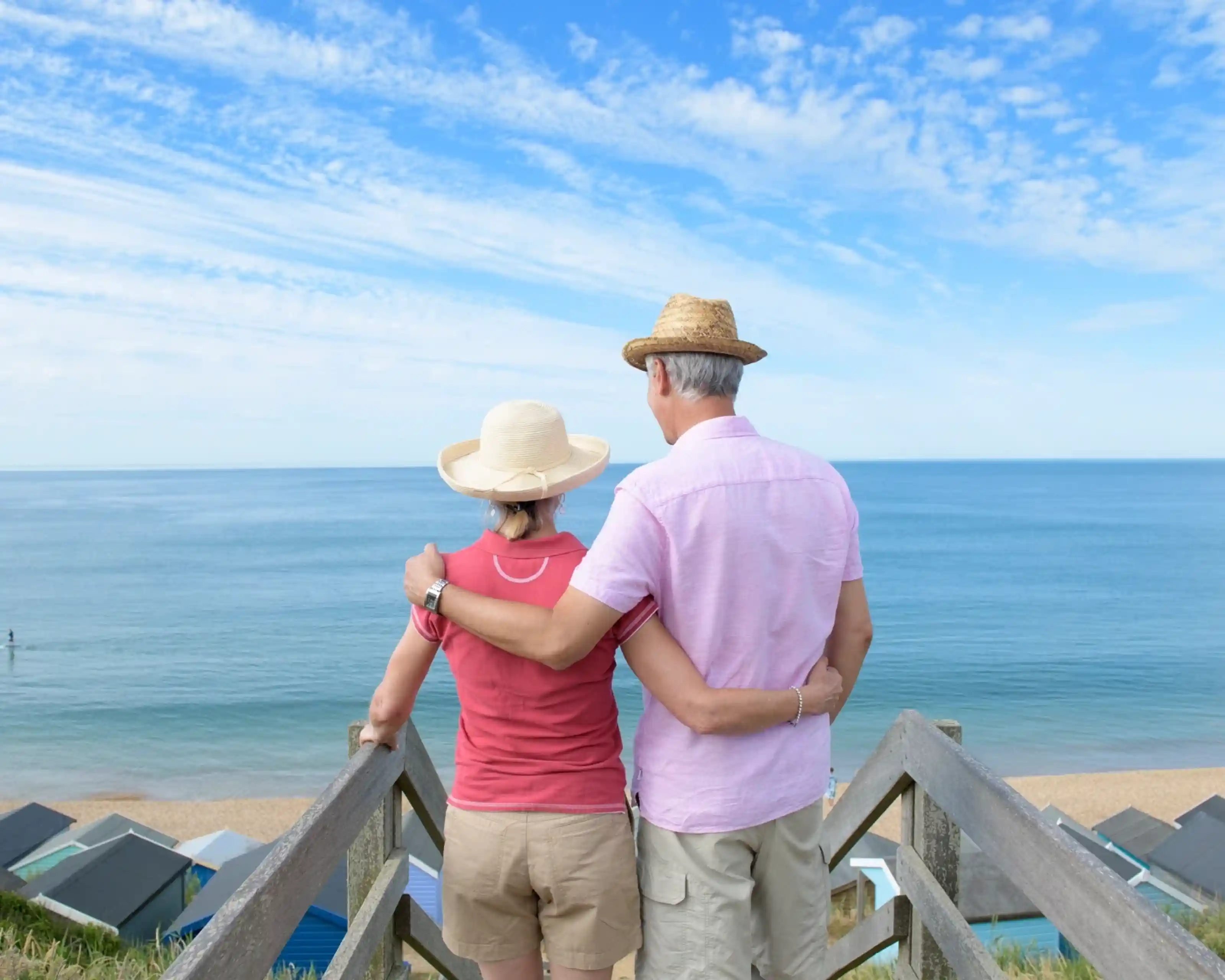 A couple stands on a wooden staircase overlooking a calm sea, with beach huts visible in the background. They are wearing summer hats and are embracing each other while enjoying the tranquil view. The sky is clear, adding to the serene atmosphere.