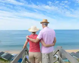 A couple stands on a wooden staircase overlooking a calm sea, with beach huts visible in the background. They are wearing summer hats and are embracing each other while enjoying the tranquil view. The sky is clear, adding to the serene atmosphere.