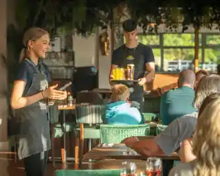 A young female server smiles while holding a notepad, standing in a bustling restaurant. In the background, a male server carries a tray with drinks, as people sit at tables enjoying their meals. The interior features lush greenery and warm lighting.
