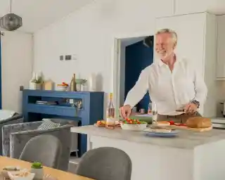 A smiling older man in a white shirt prepares food on a kitchen island, with a bottle of rosé wine and a bowl of salad. The modern kitchen features soft colors and stylish decor.