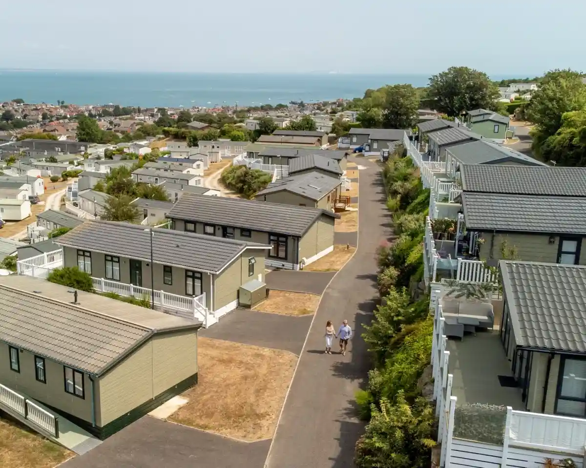 Aerial view of a coastal residential area with modular homes along a winding road. Two people are walking together on the pathway, surrounded by greenery and views of the ocean in the background.