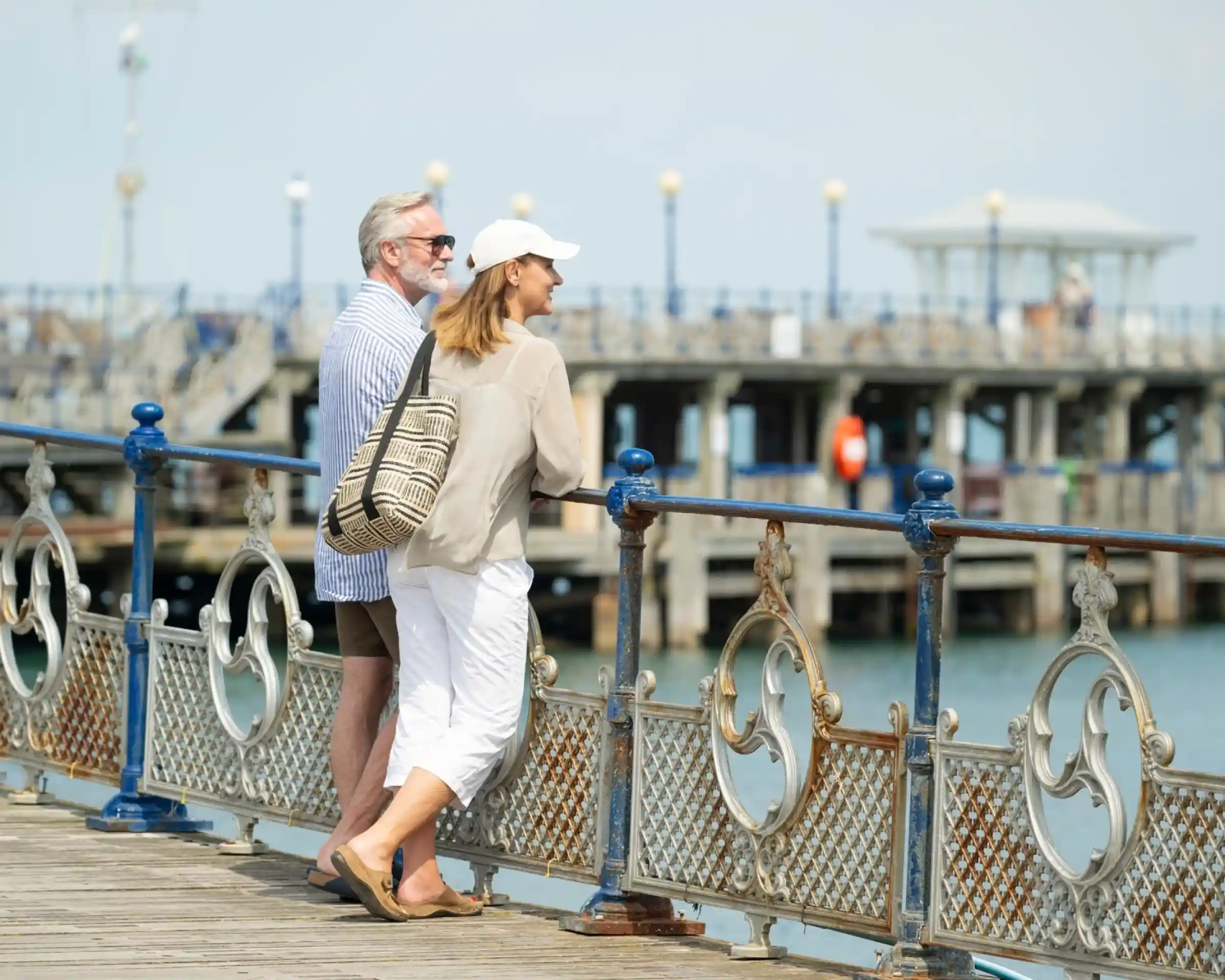 A couple stands on a wooden pier, smiling and enjoying the view of the water. The man has gray hair and wears sunglasses with a striped shirt and shorts. The woman, wearing a cap and a light blouse, leans against the railing, holding a striped bag. A pier with buildings and lamps is visible in the background.