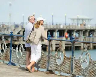 A couple stands on a wooden pier, smiling and enjoying the view of the water. The man has gray hair and wears sunglasses with a striped shirt and shorts. The woman, wearing a cap and a light blouse, leans against the railing, holding a striped bag. A pier with buildings and lamps is visible in the background.