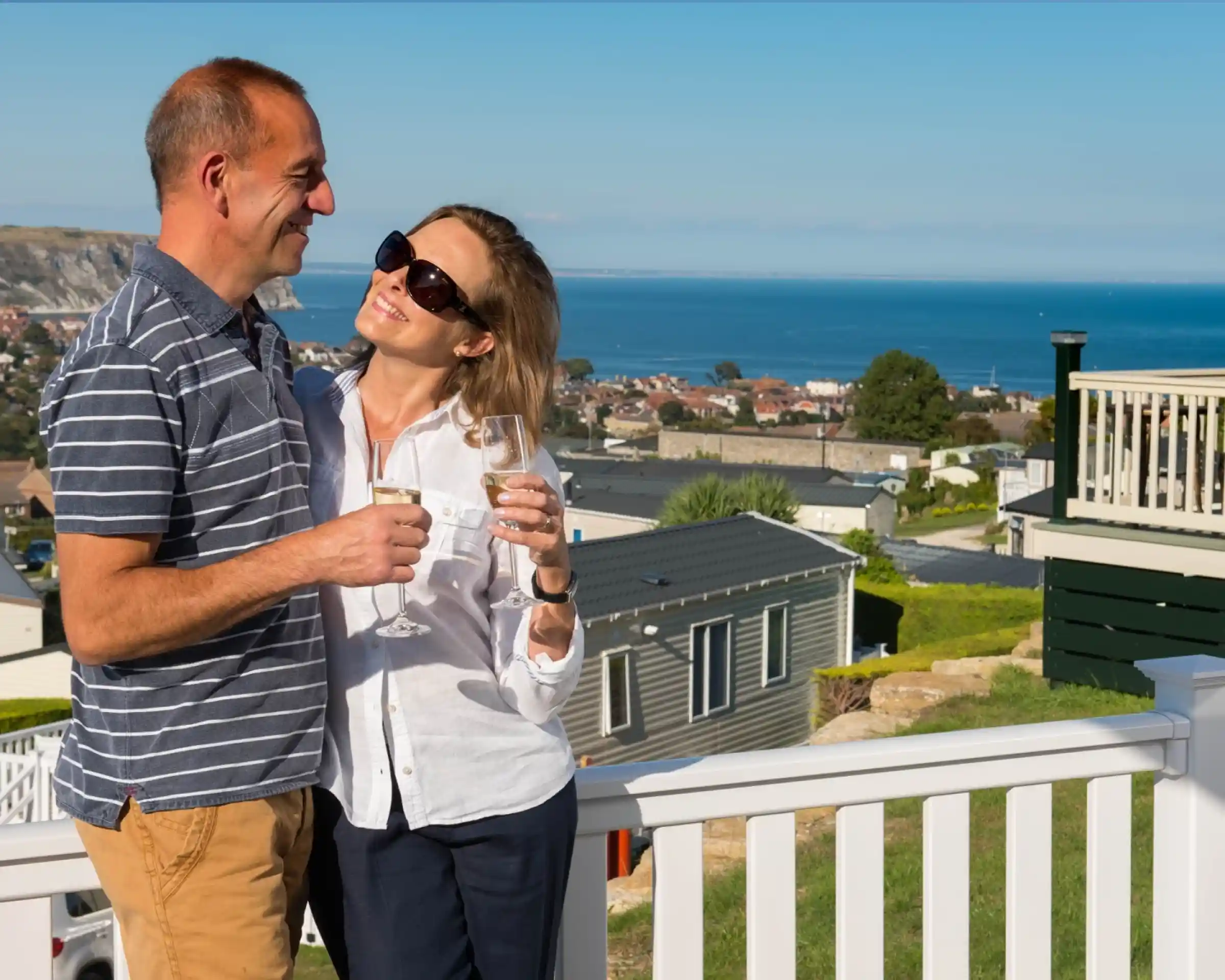 A couple stands on a balcony overlooking a scenic view of the ocean and coastline. They are smiling and holding glasses of sparkling drink, dressed casually in a striped shirt and white blouse with sunglasses on.