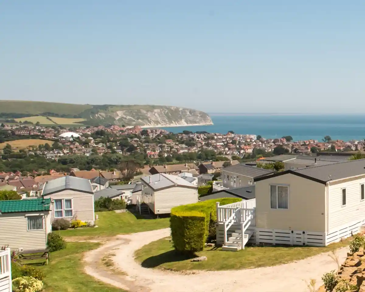 A scenic view overlooking a coastal town with a cluster of caravans in the foreground. Rolling hills and cliffs rise in the background, meeting a clear blue sea under a bright sky.
