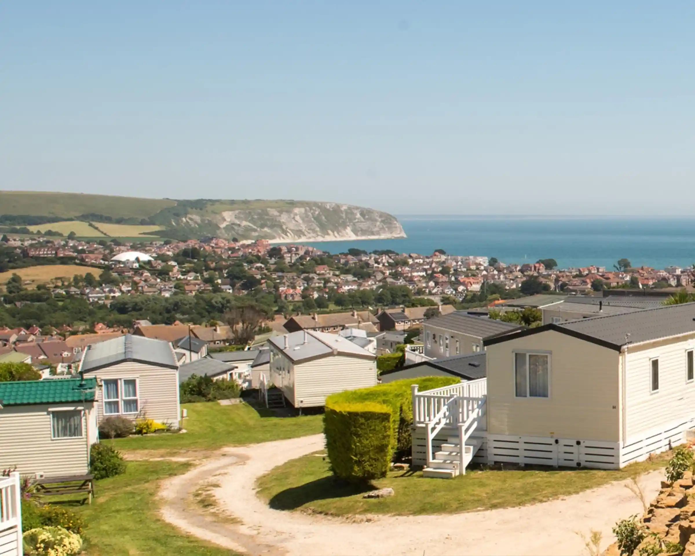 A scenic view overlooking a coastal town with a cluster of caravans in the foreground. Rolling hills and cliffs rise in the background, meeting a clear blue sea under a bright sky.