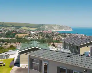 A panoramic view of a coastal landscape featuring colorful mobile homes in the foreground, lush green hills in the background, and a clear blue sky above. The sea is visible along with a distant cliffside.