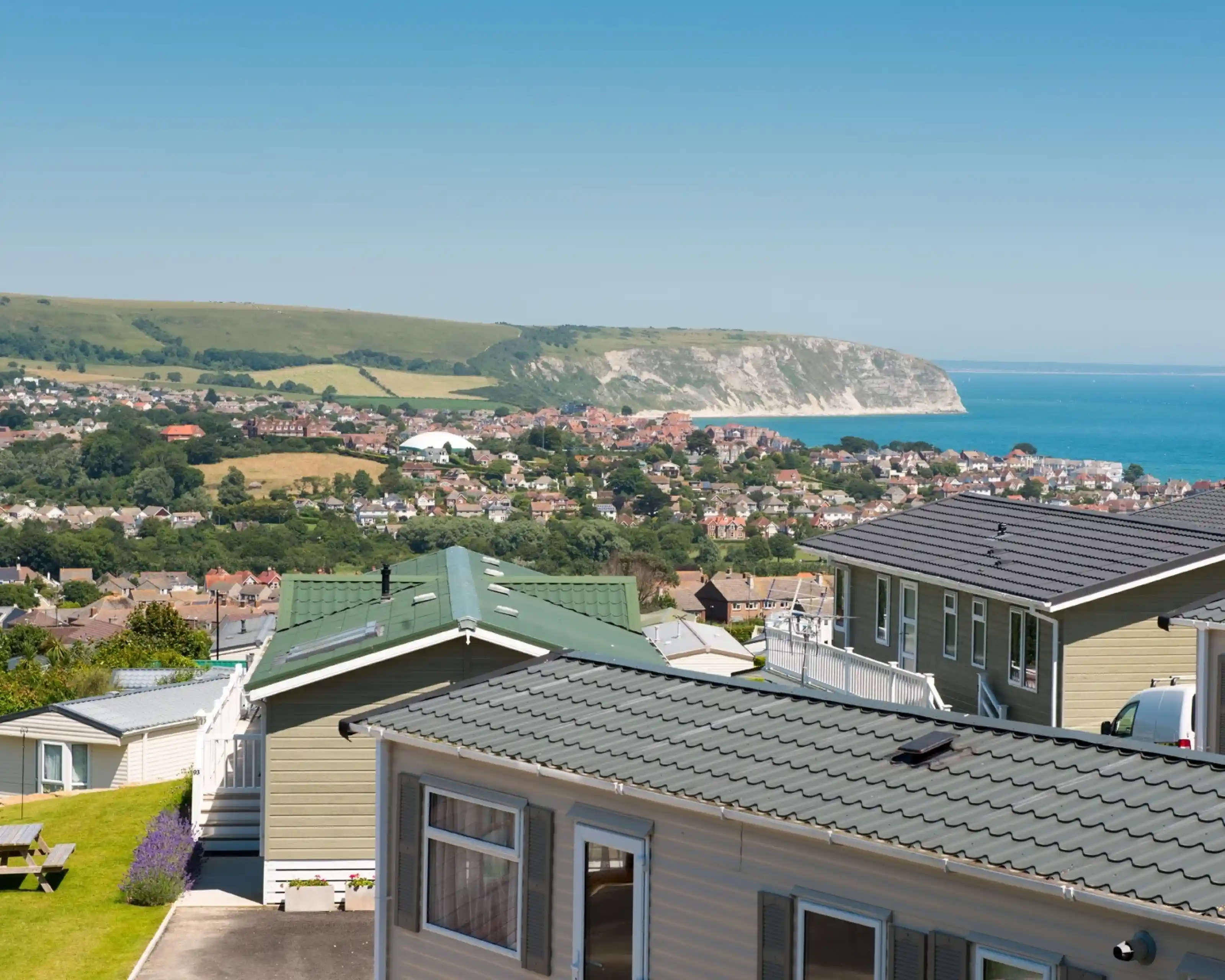 A panoramic view of a coastal landscape featuring colorful mobile homes in the foreground, lush green hills in the background, and a clear blue sky above. The sea is visible along with a distant cliffside.