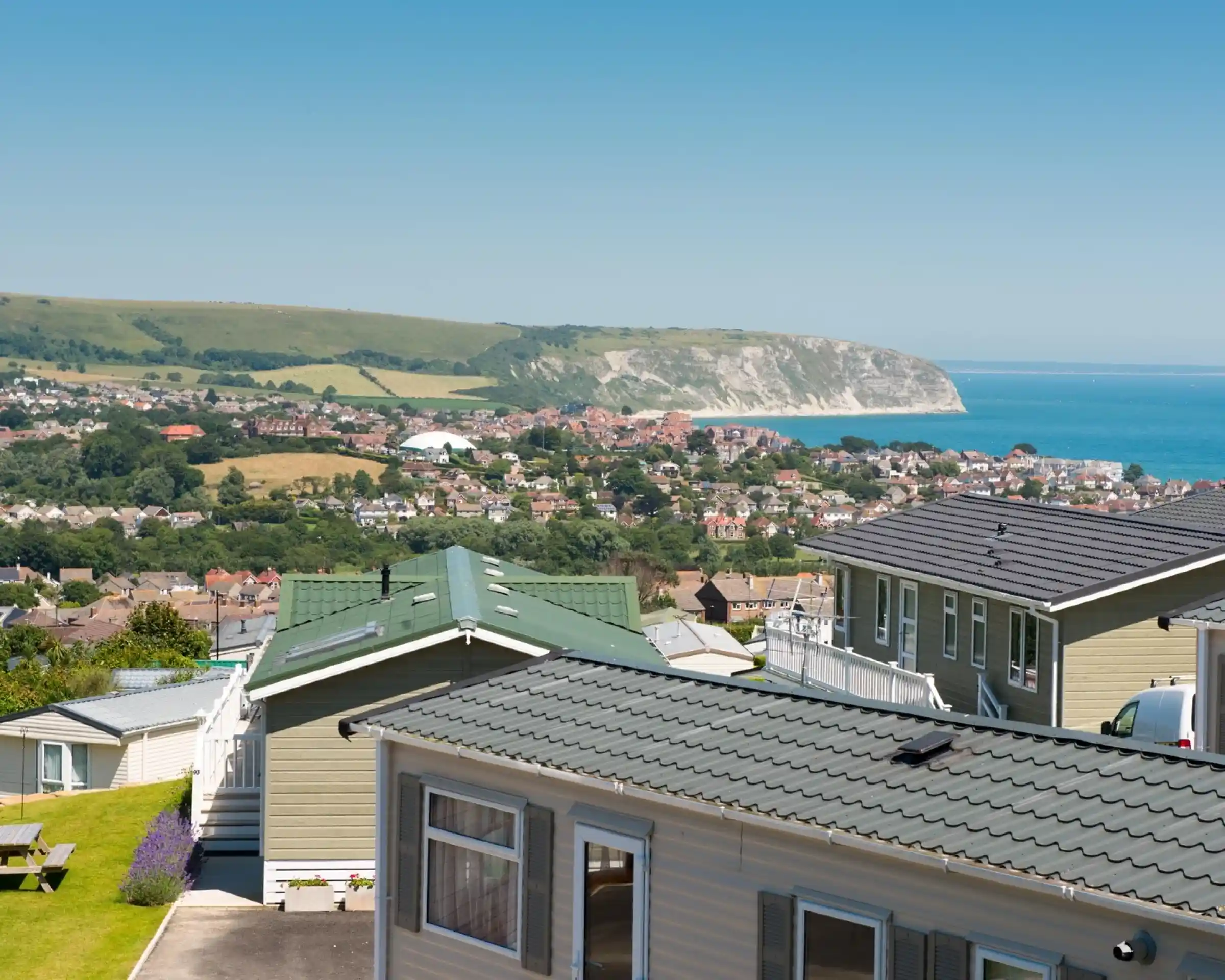 A panoramic view of a coastal landscape featuring colorful mobile homes in the foreground, lush green hills in the background, and a clear blue sky above. The sea is visible along with a distant cliffside.
