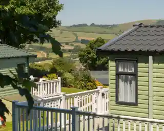 A green holiday home with a black roof is situated on a hillside. The surrounding landscape features rolling hills, lush greenery, and colorful flowers. A white wooden railing leads to the entrance of the home.