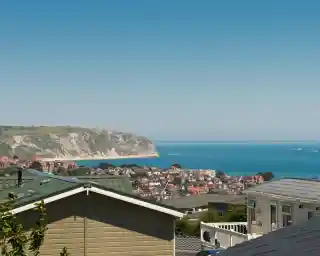 A panoramic view of a coastal town with colorful houses nestled among green hills, overlooking a blue sea and cliffs under a clear blue sky.
