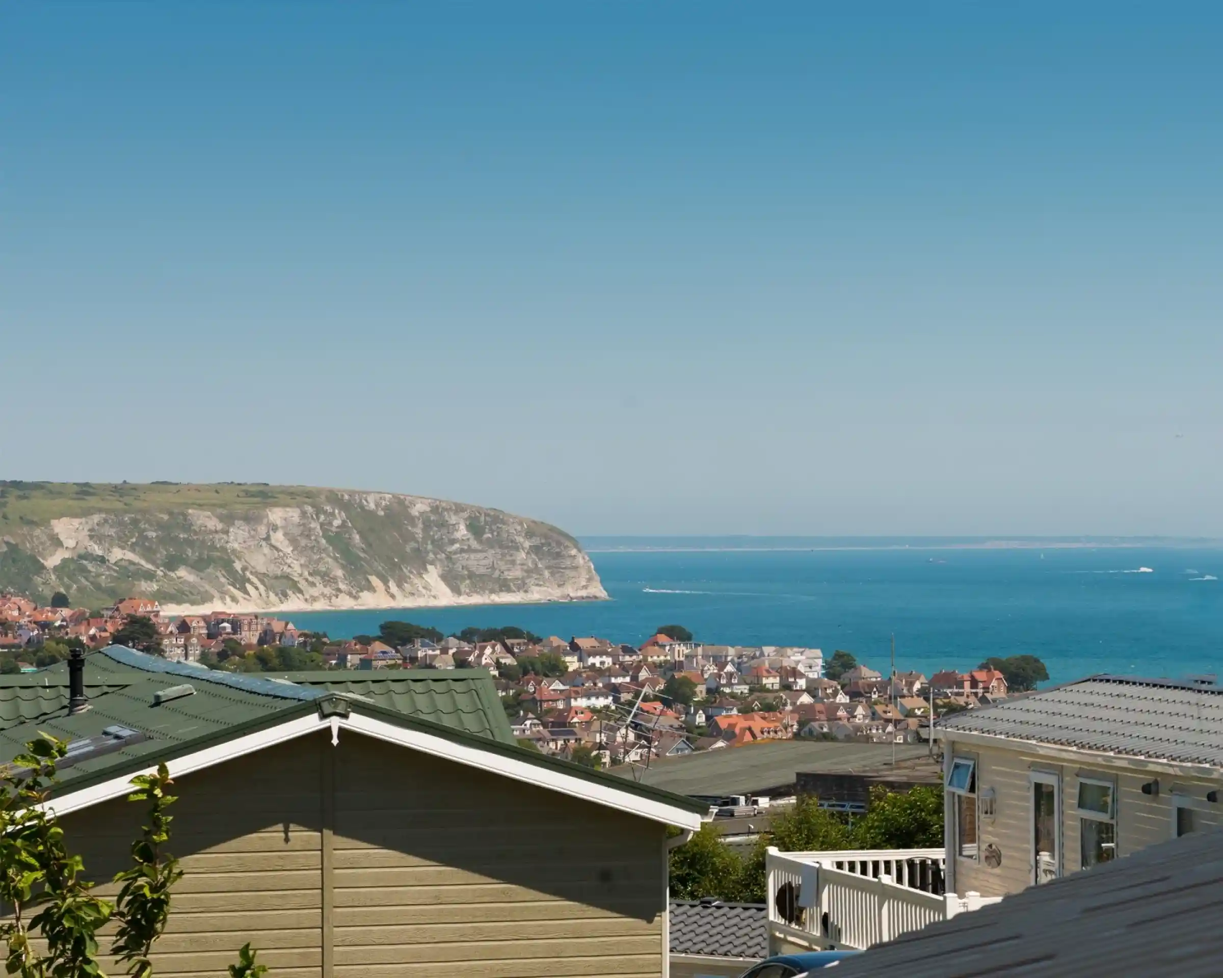 A panoramic view of a coastal town with colorful houses nestled among green hills, overlooking a blue sea and cliffs under a clear blue sky.