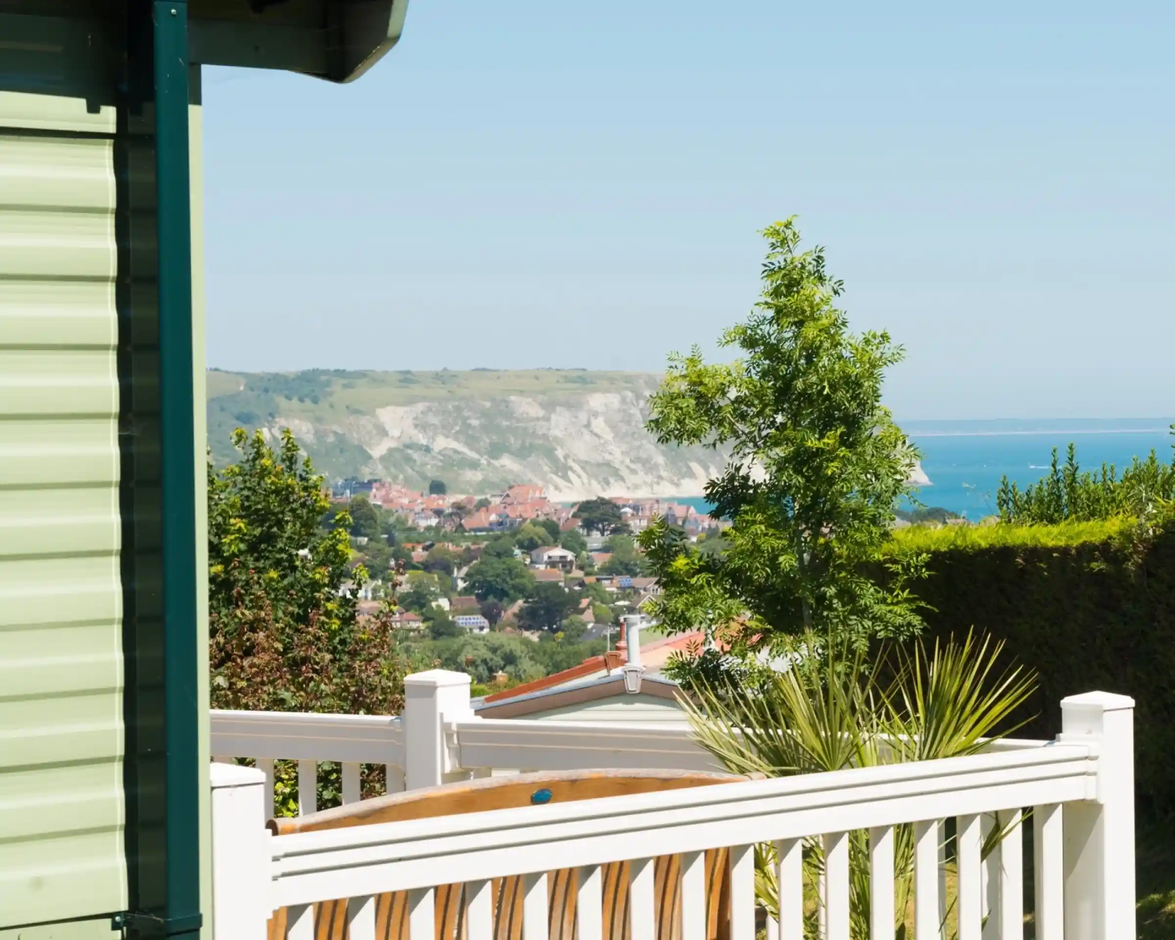 A view of a coastal town with white cliffs in the background. Green trees and plants are visible in the foreground, along with a wooden seating area. The sky is clear and blue.