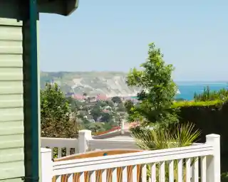 A view of a coastal town with white cliffs in the background. Green trees and plants are visible in the foreground, along with a wooden seating area. The sky is clear and blue.