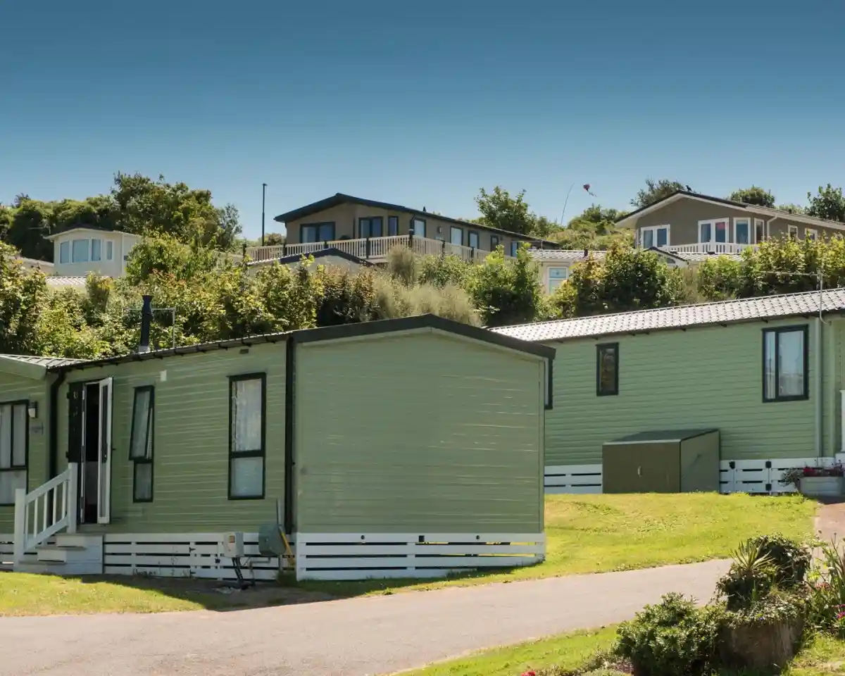 Green modular homes are lined up in a scenic area, with trees in the background. The sky is clear and blue, and some homes are positioned on a hill. A paved path runs in front of the houses.