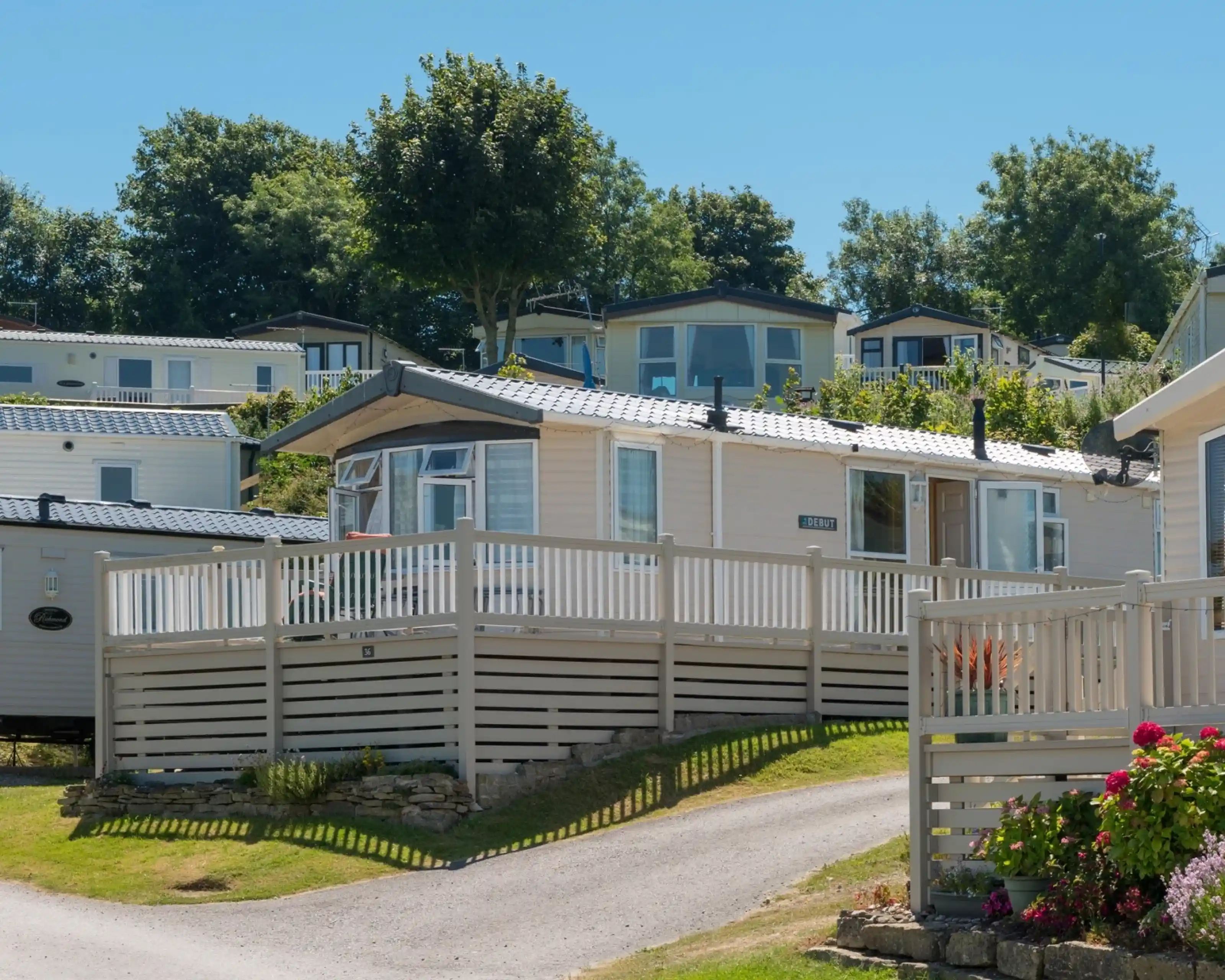 A holiday home in a trailer park, featuring a wooden deck and a picturesque outdoor setting. Surrounding greenery includes trees and shrubs, with additional homes visible on a hillside in the background. Clear blue sky enhances the inviting atmosphere.