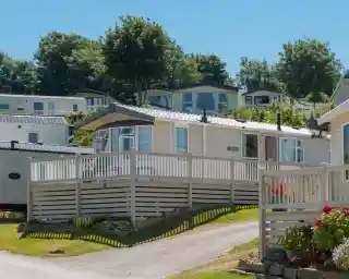 A holiday home in a trailer park, featuring a wooden deck and a picturesque outdoor setting. Surrounding greenery includes trees and shrubs, with additional homes visible on a hillside in the background. Clear blue sky enhances the inviting atmosphere.