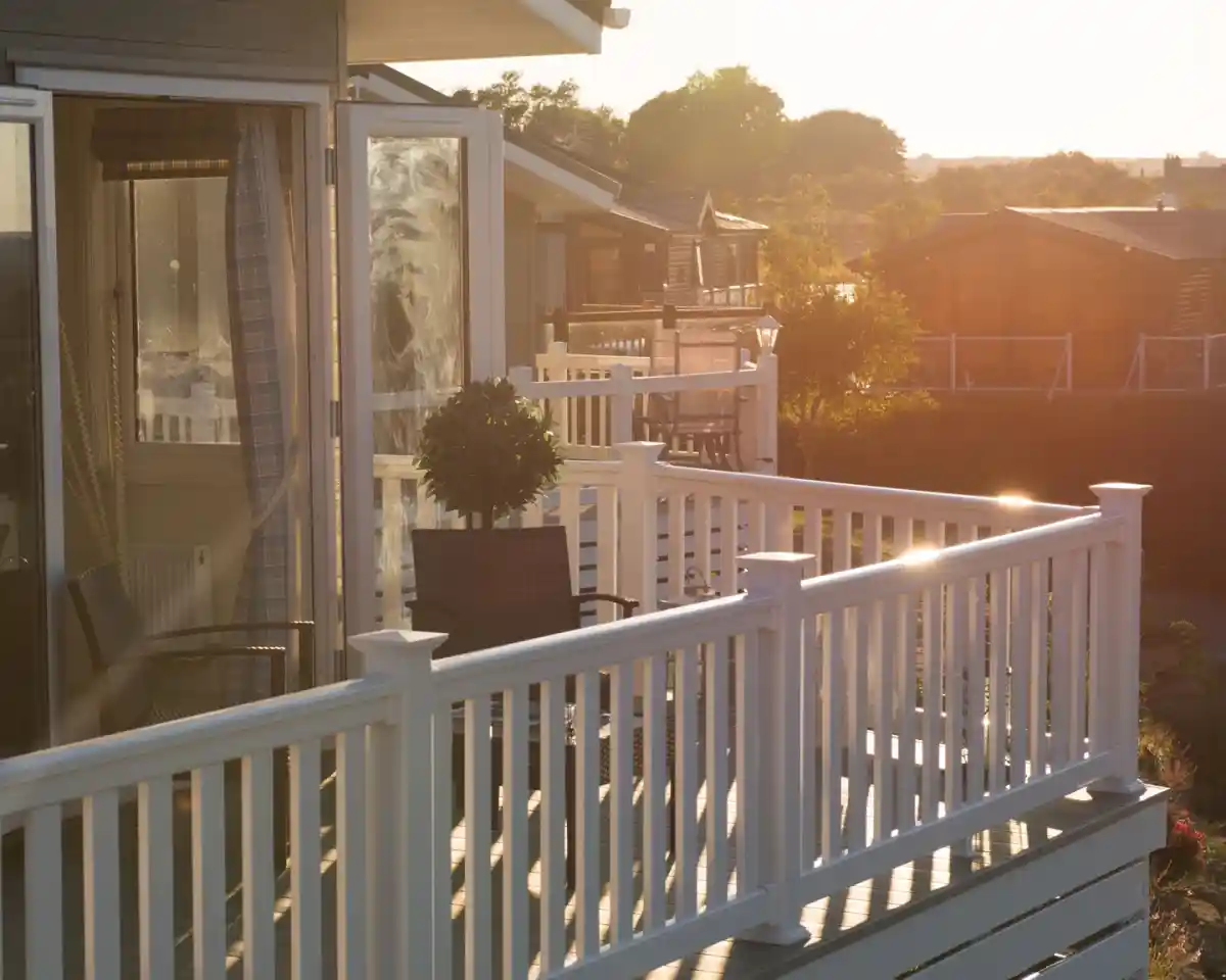 A sunlit patio with white railings overlooks a tranquil scene of wooden cottages in the background. A potted plant is placed on the table, and the warm glow of sunset creates a serene atmosphere.