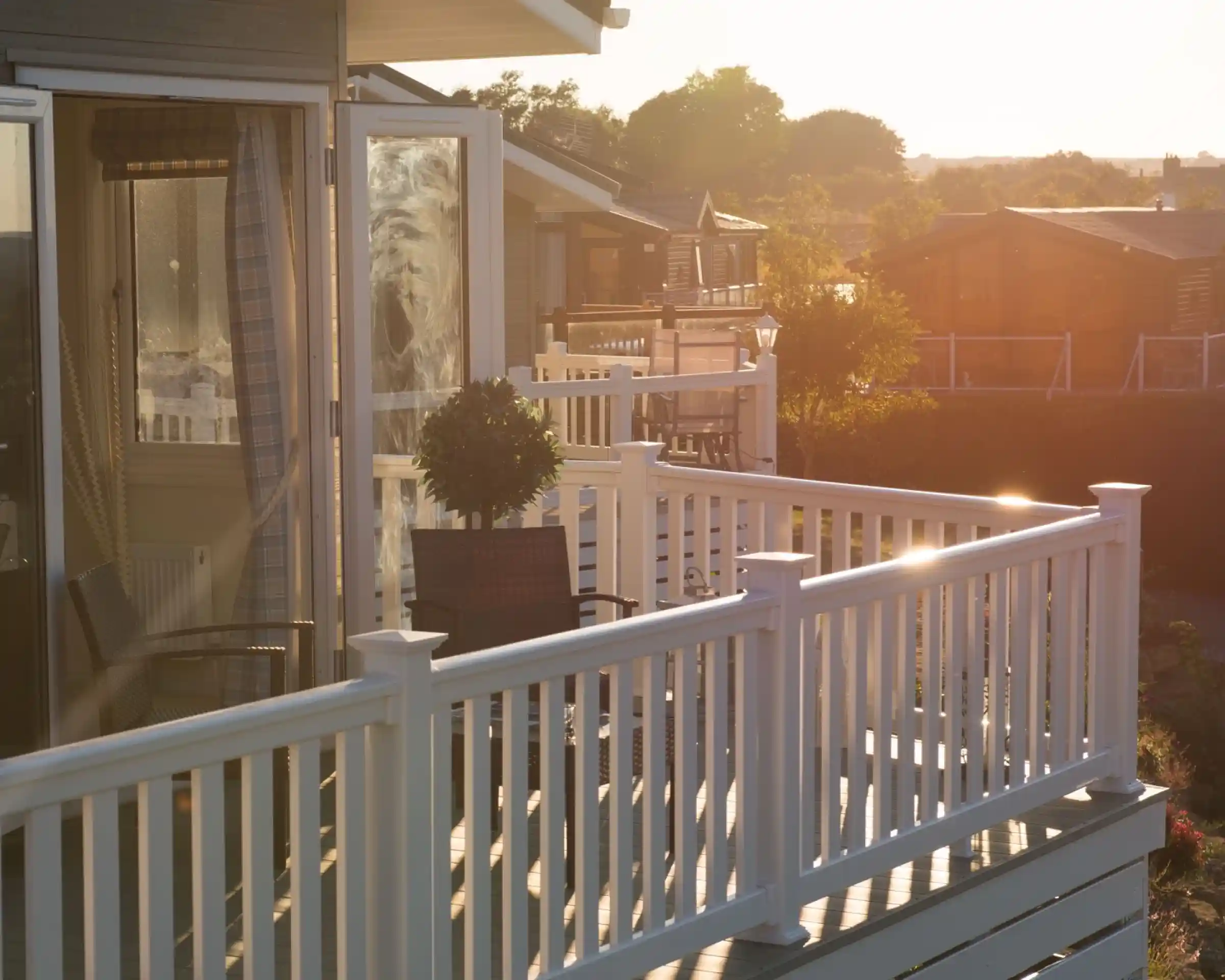 A sunlit patio with white railings overlooks a tranquil scene of wooden cottages in the background. A potted plant is placed on the table, and the warm glow of sunset creates a serene atmosphere.