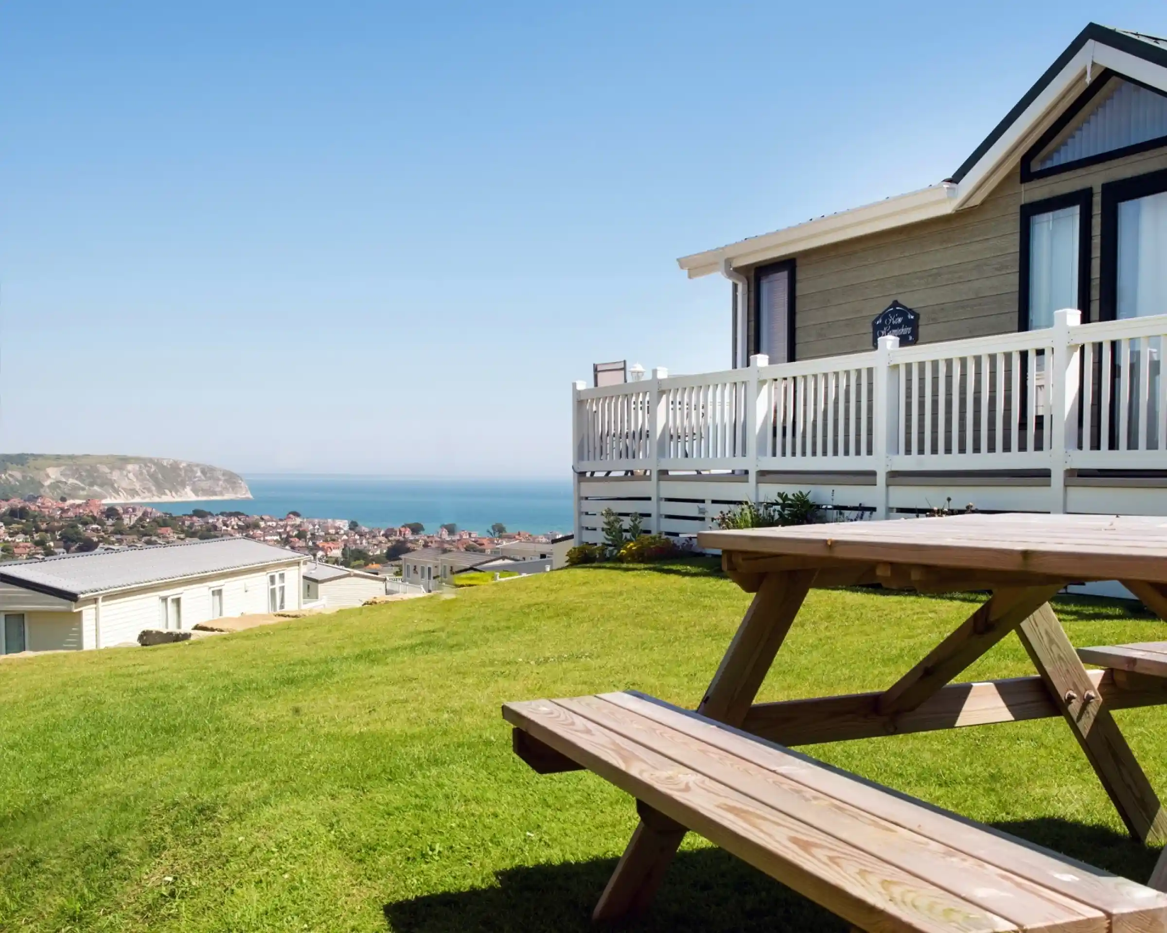 A wooden picnic table sits on a grassy area in the foreground, overlooking a coastal view. A modern home with a porch is visible to the right, framed by clear blue skies and a distant shoreline in the background.