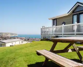 A wooden picnic table sits on a grassy area in the foreground, overlooking a coastal view. A modern home with a porch is visible to the right, framed by clear blue skies and a distant shoreline in the background.