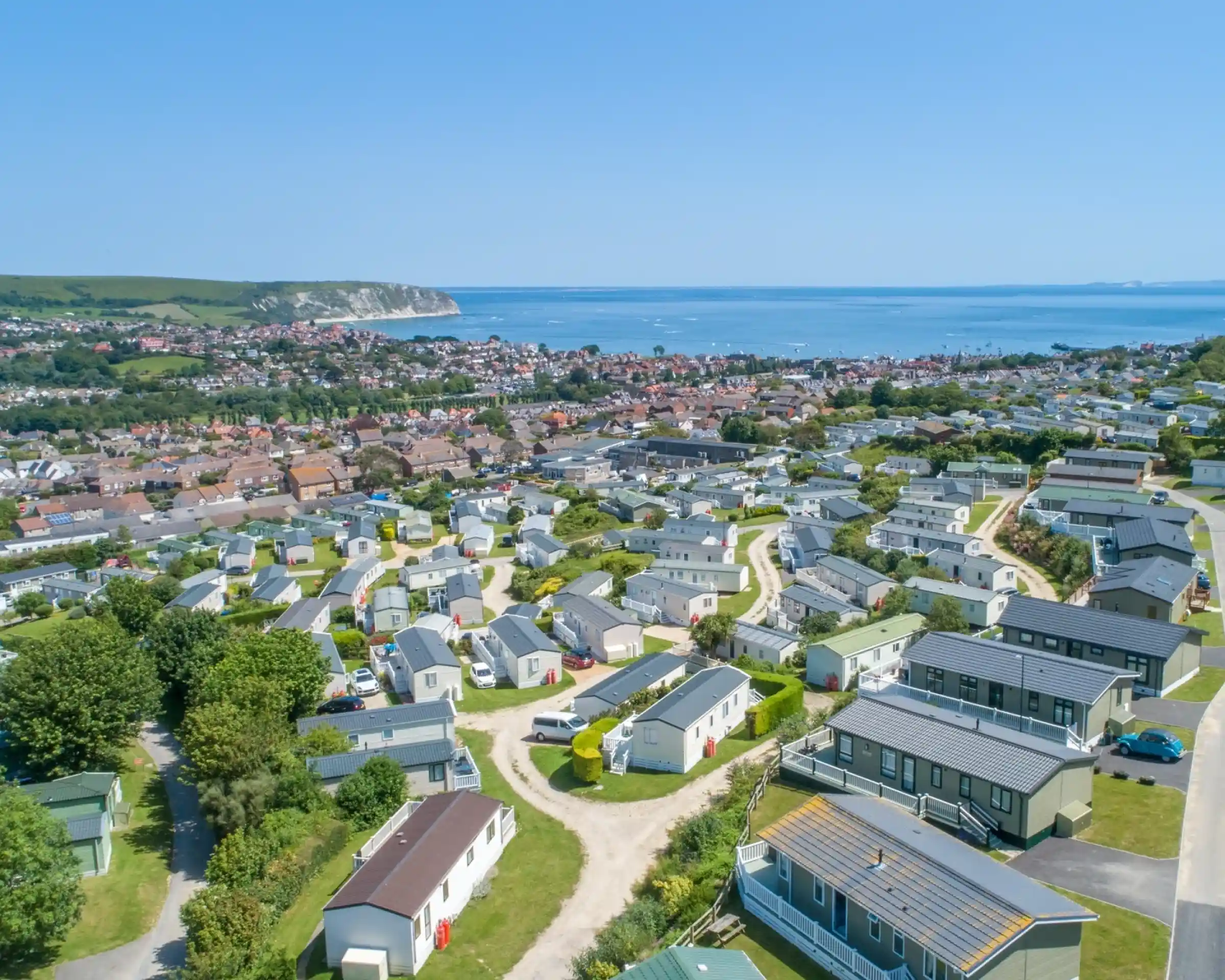 Aerial view of a coastal town featuring rows of holiday homes and caravans, surrounded by greenery and overlooking the ocean. Cliffs and hills are visible in the background.