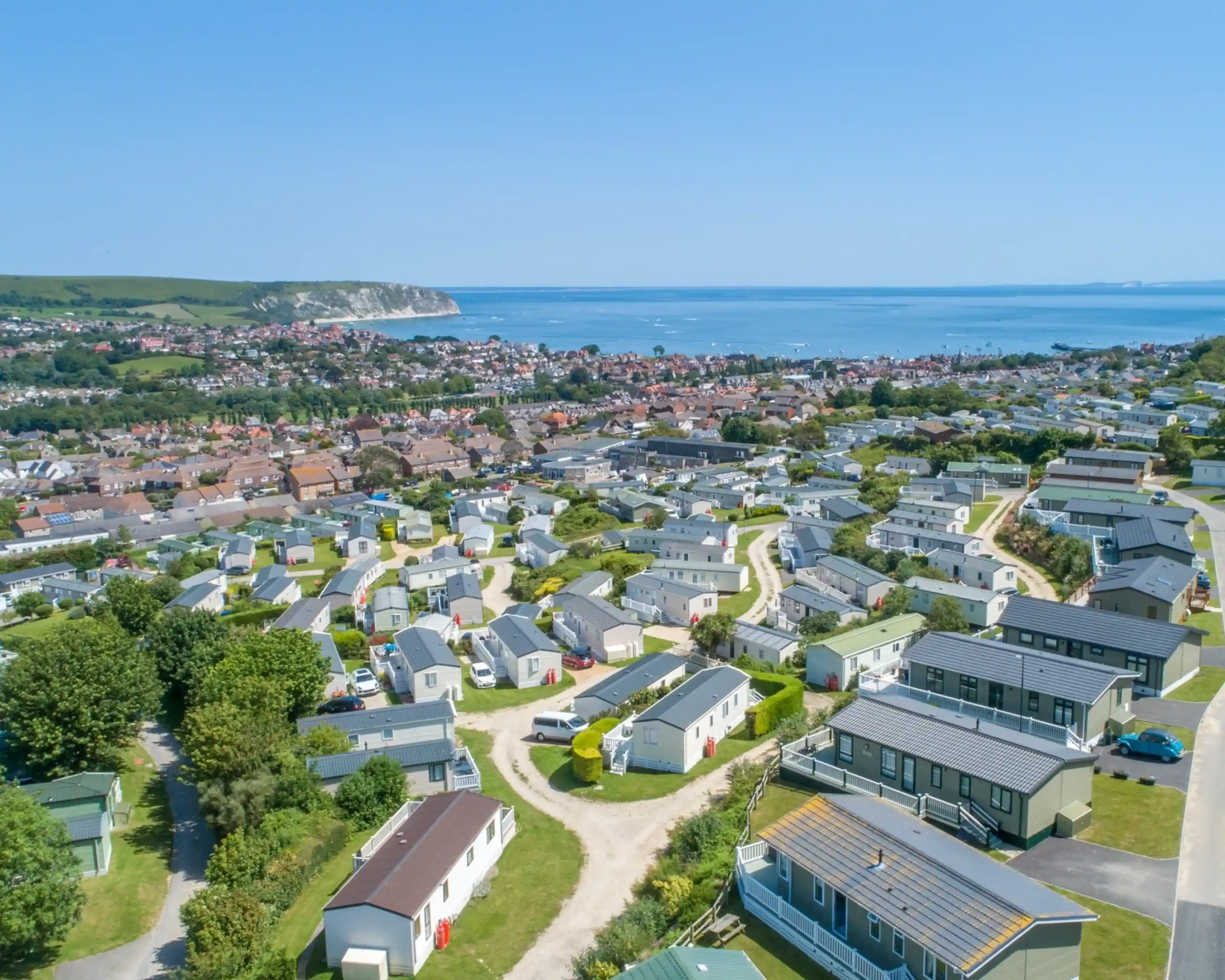 Aerial view of a coastal town featuring rows of holiday homes and caravans, surrounded by greenery and overlooking the ocean. Cliffs and hills are visible in the background.