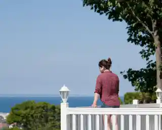 A woman stands on a balcony railing, gazing out at a serene ocean view. She is wearing a red plaid shirt and shorts, with her hair tied back. Lush greenery surrounds her near the railing, under a clear blue sky.