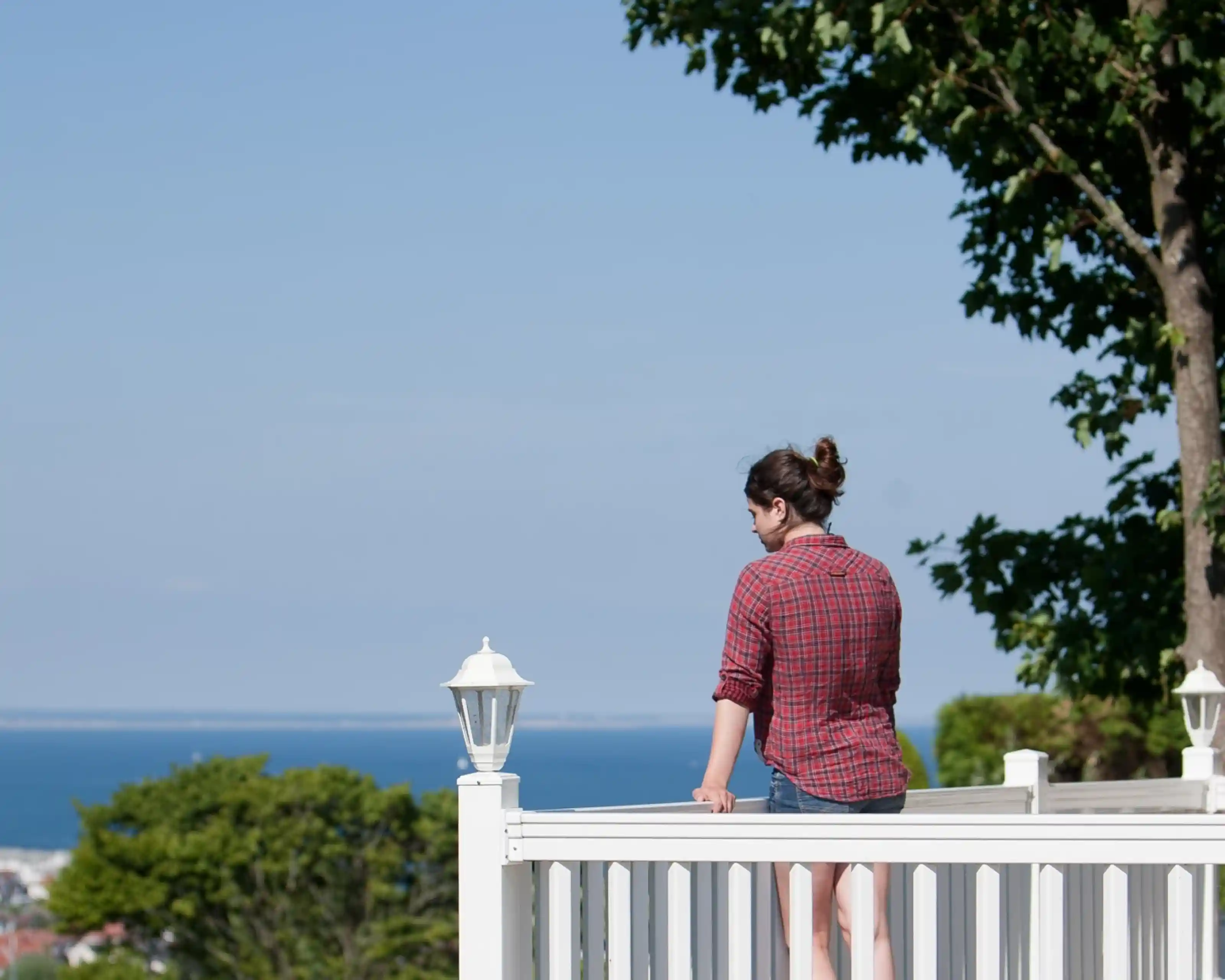 A woman stands on a balcony railing, gazing out at a serene ocean view. She is wearing a red plaid shirt and shorts, with her hair tied back. Lush greenery surrounds her near the railing, under a clear blue sky.