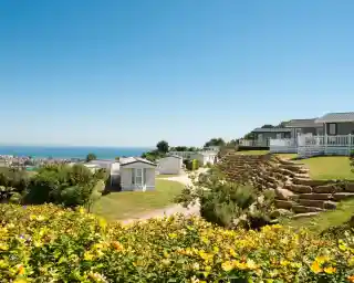 A scenic view of a coastal landscape featuring several holiday cabins on a hillside. Flowering yellow plants in the foreground lead down to a pathway, with the ocean visible in the background under a clear blue sky.