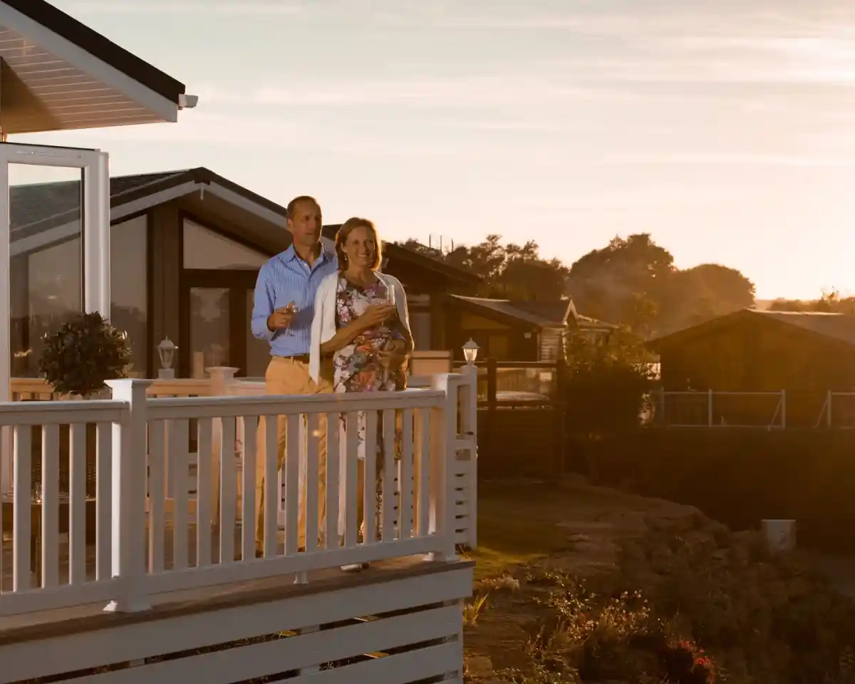 A couple stands on a deck overlooking a scenic landscape during sunset, holding drinks and smiling. Cozy cabins are in the background, surrounded by greenery.