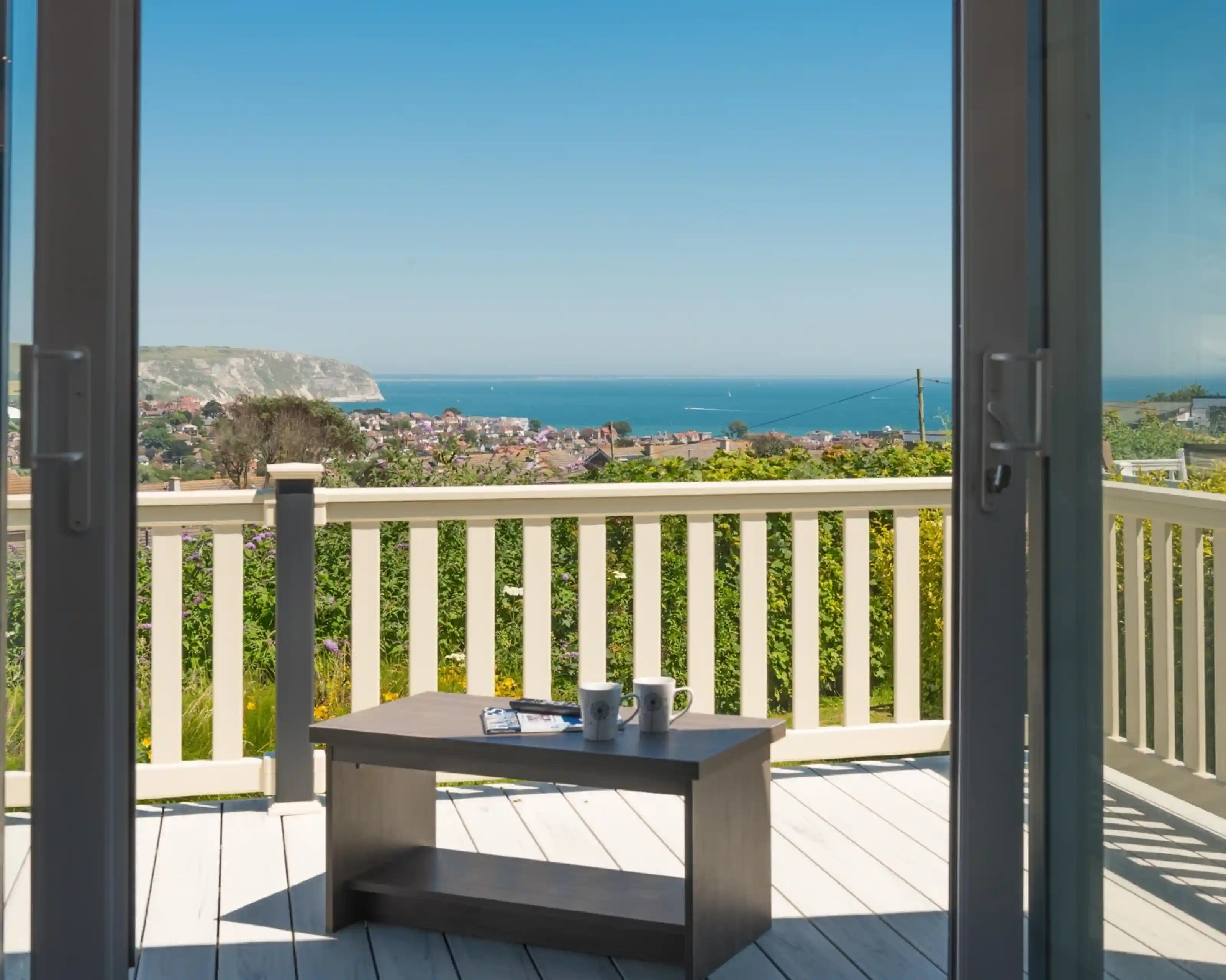 A view from a balcony overlooking a coastal landscape, featuring the ocean and distant cliffs. A small wooden table with two mugs sits in the foreground, framed by sliding glass doors. Bright blue sky and clear weather enhance the scenic backdrop.