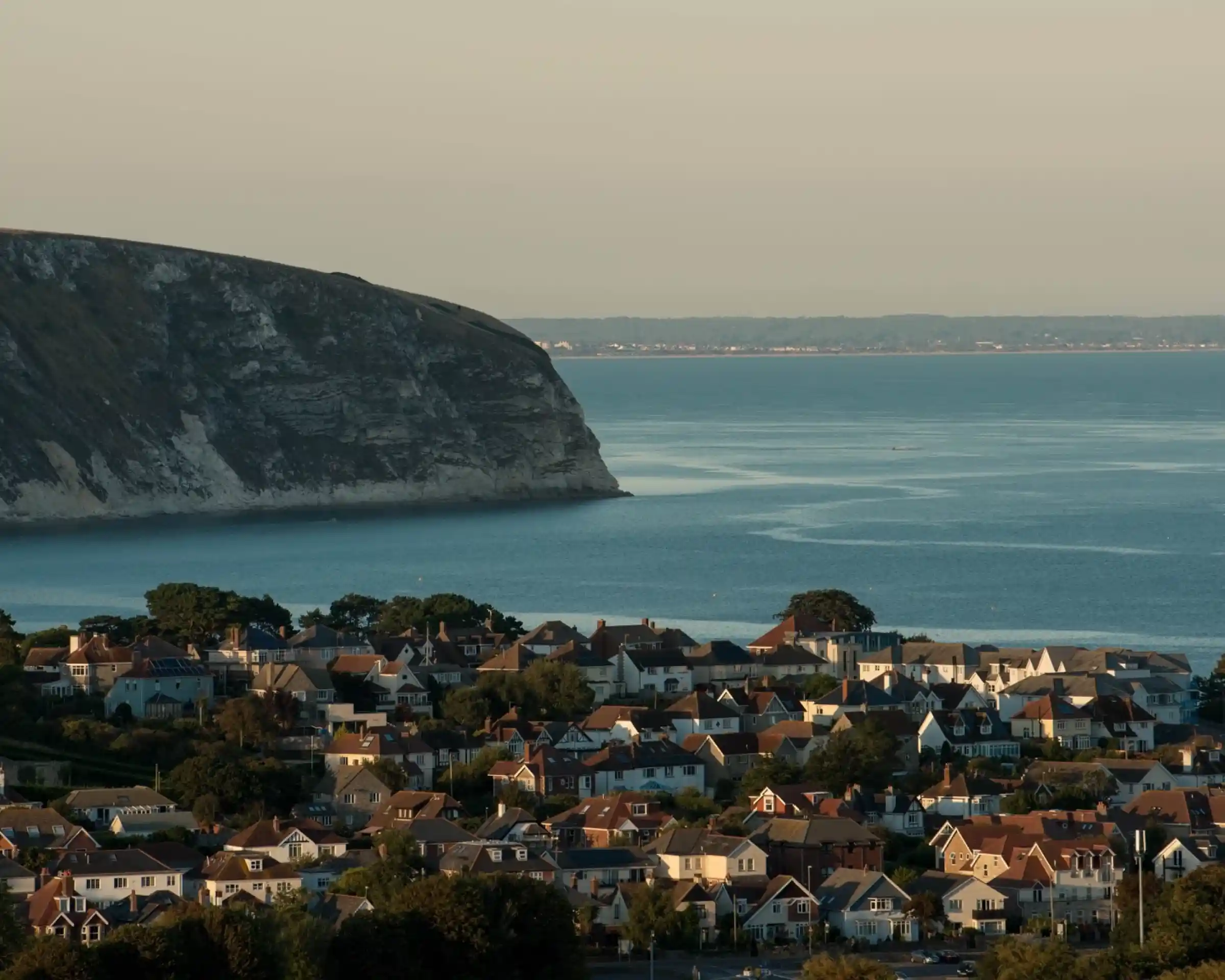 A coastal view featuring a hillside of residential homes with varying architectural styles. In the background, a large cliff towers above the calm sea, under a clear sky.