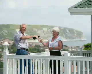 A man pours champagne into a glass held by a woman on a balcony, with a coastal cliff and ocean in the background. They appear joyful and celebratory.
