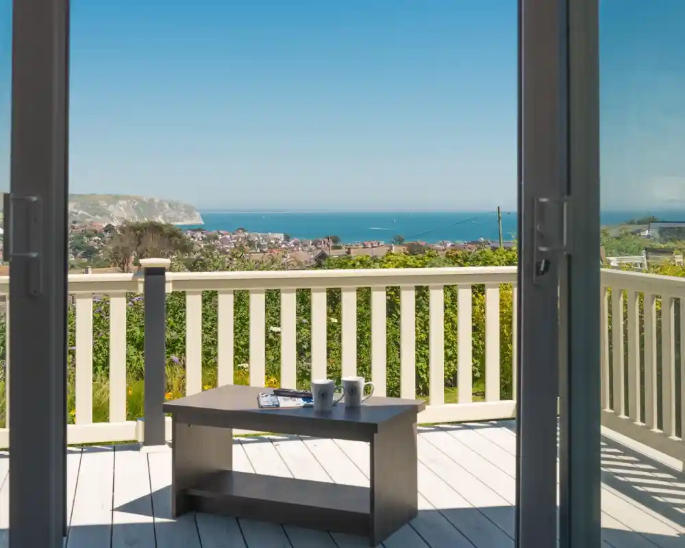 A wooden deck overlooks a coastal view with a small table holding two cups. The ocean and distant hills are visible under a clear blue sky.