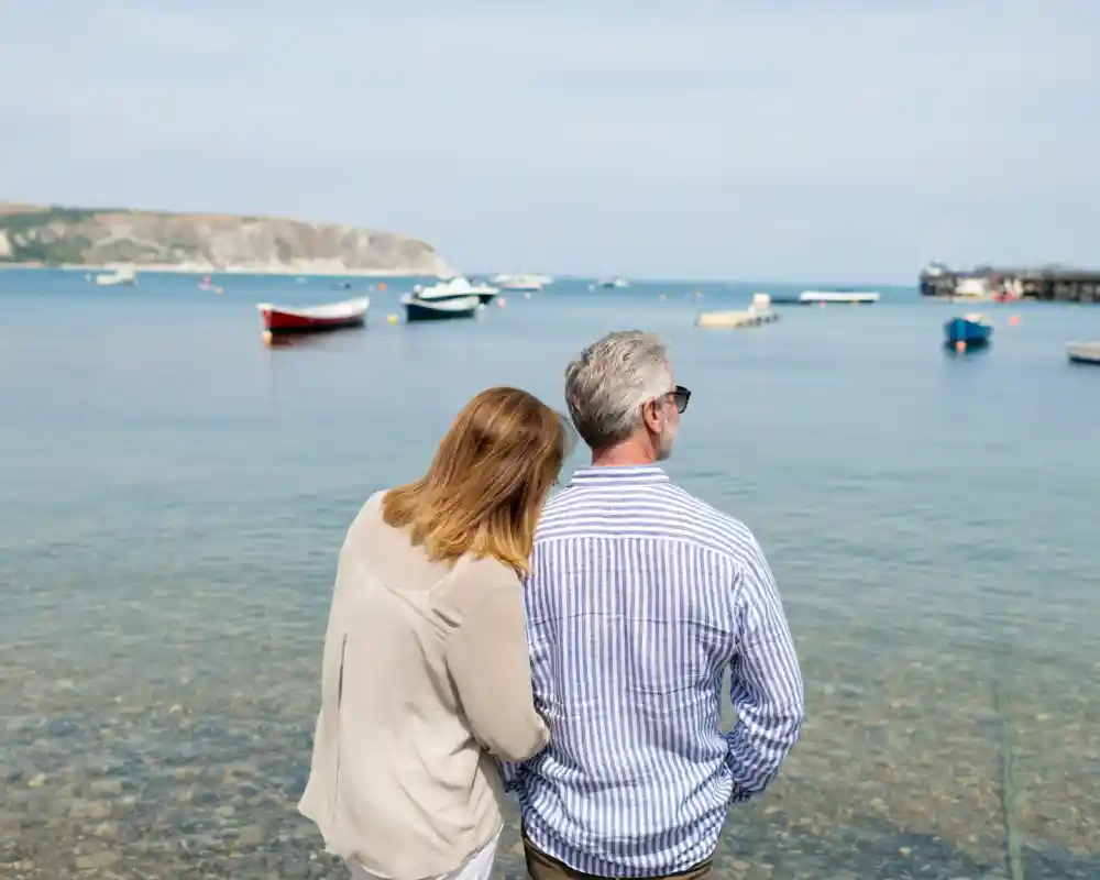 A middle-aged couple stands together on the shoreline, with the woman leaning her head against the man's back. They face a tranquil sea dotted with small boats, while a pier is visible in the distance. The sky is clear, and the water is calm, reflecting the surrounding scenery.