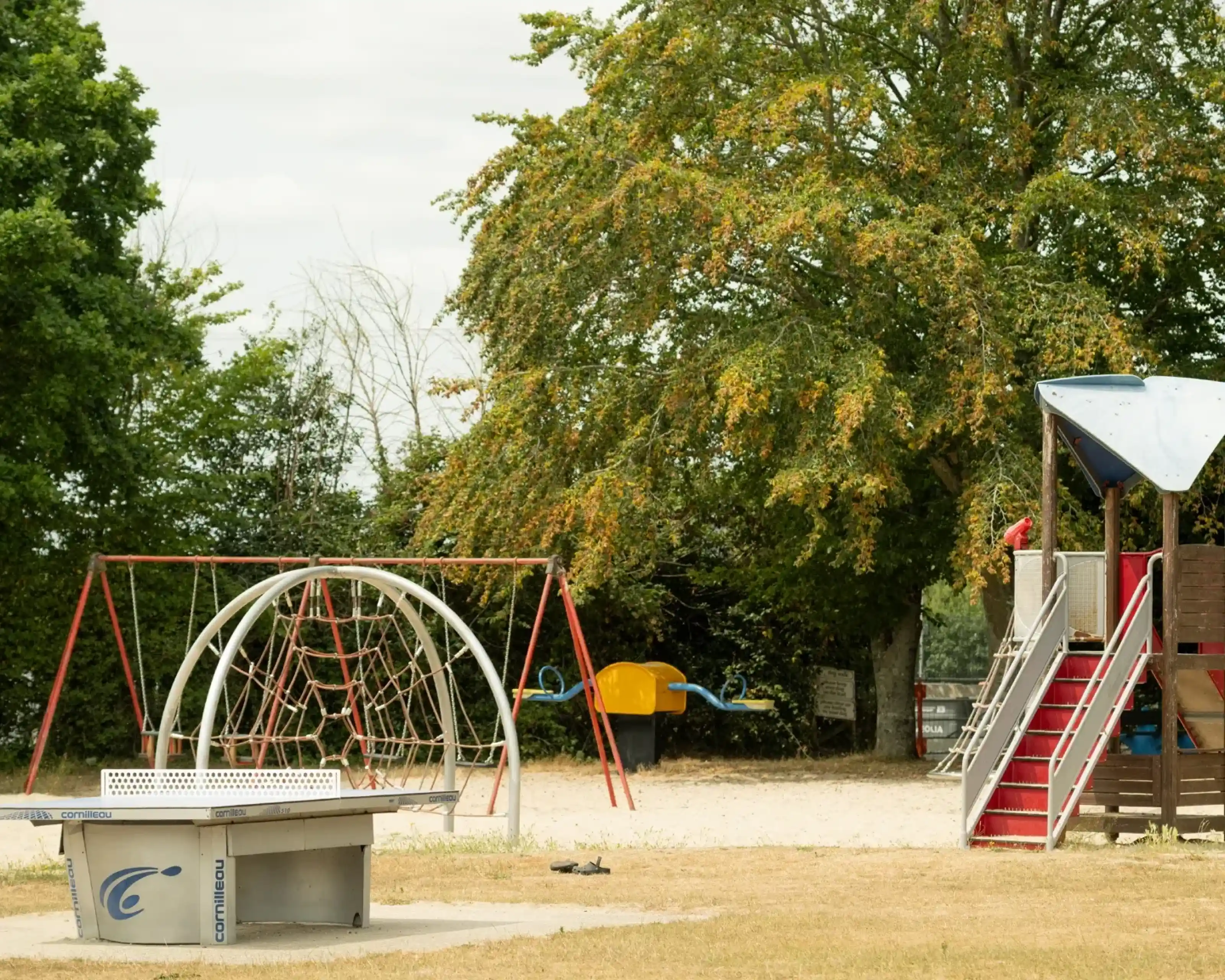 A playground features a climbing frame, swings, and a slide, surrounded by trees and a grassy area. It appears to be a calm day with a few clouds in the sky.
