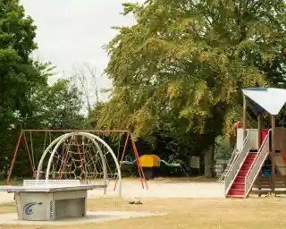 A playground features a climbing frame, swings, and a slide, surrounded by trees and a grassy area. It appears to be a calm day with a few clouds in the sky.