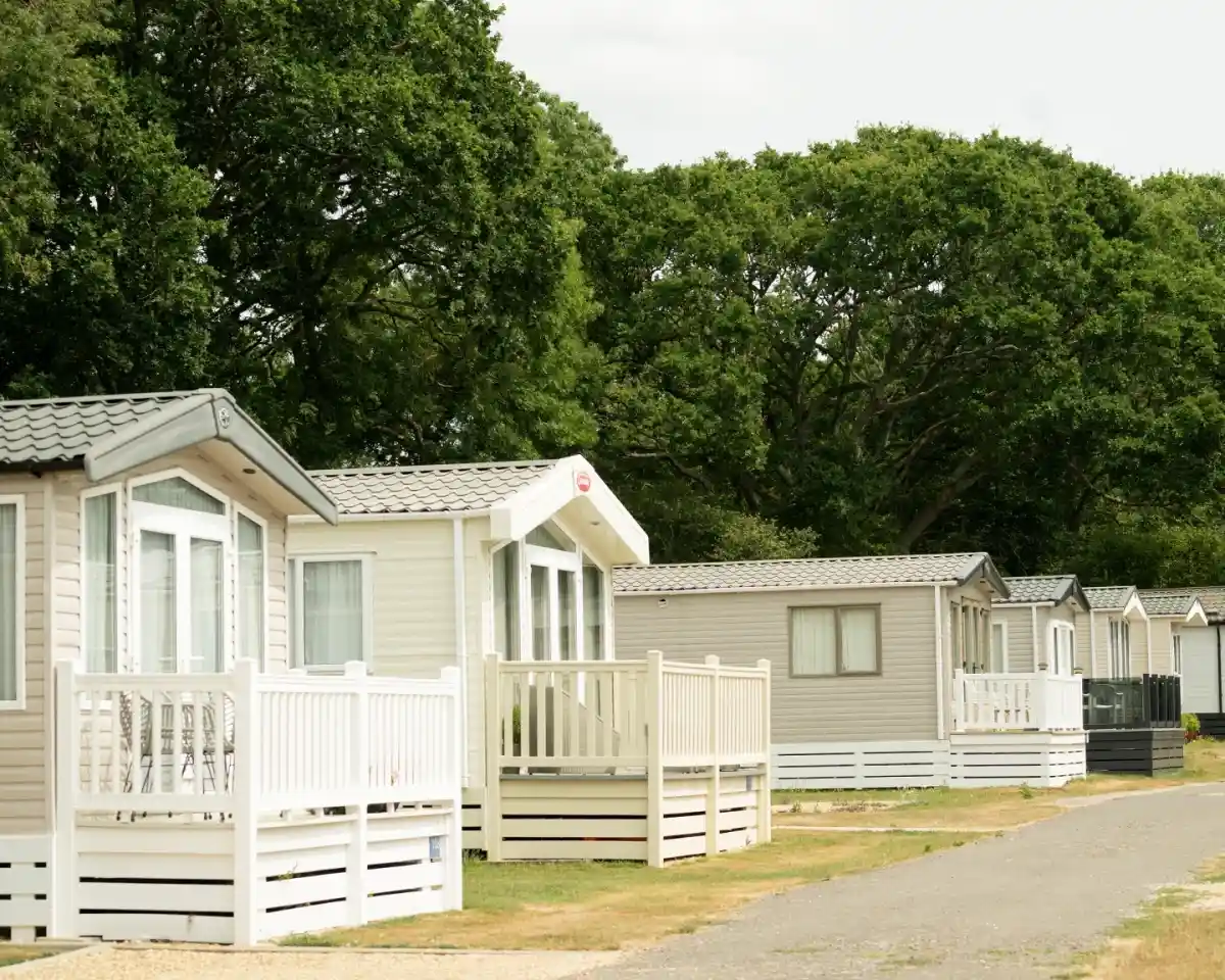 Row of modern static caravans with white porches, surrounded by greenery and trees. The sky is partly cloudy, indicating a calm day.