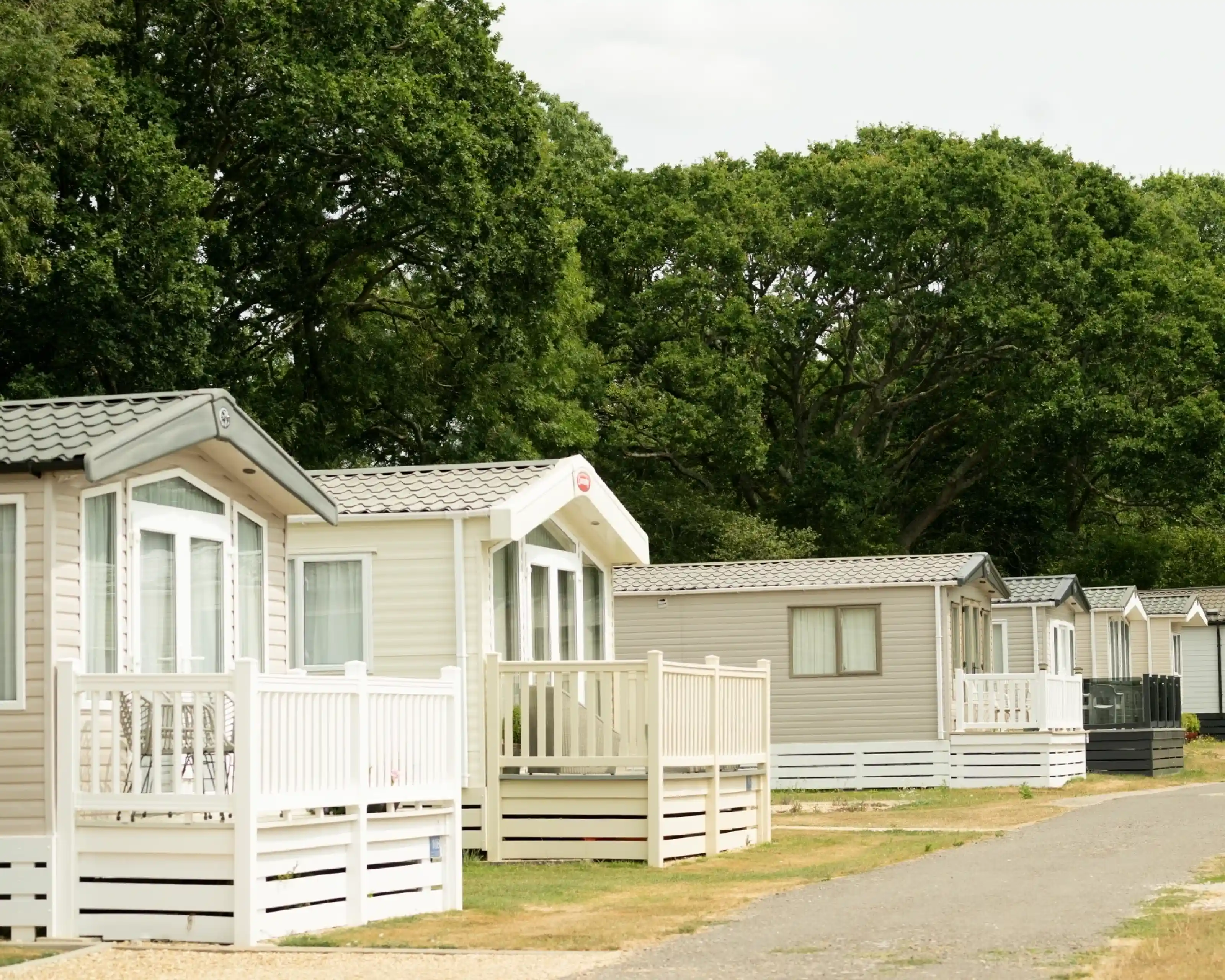 Row of modern static caravans with white porches, surrounded by greenery and trees. The sky is partly cloudy, indicating a calm day.