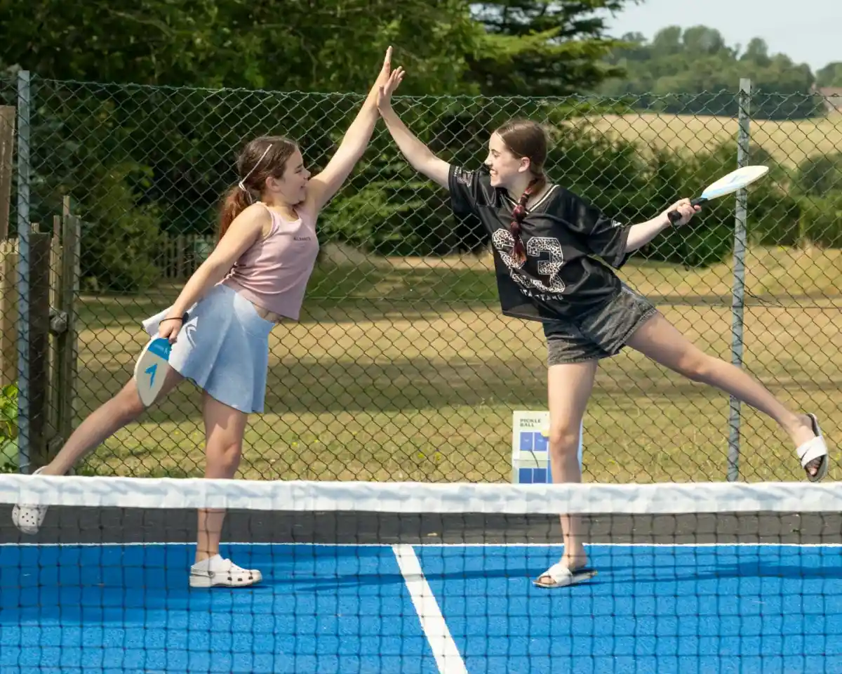 Two girls play tennis on a blue court, striking a pose as they hold their rackets. One girl is wearing a pink top and a blue skirt, while the other is dressed in a black oversized shirt and shorts. They are both smiling and appear to be having fun. A fence and grassy area are visible in the background.