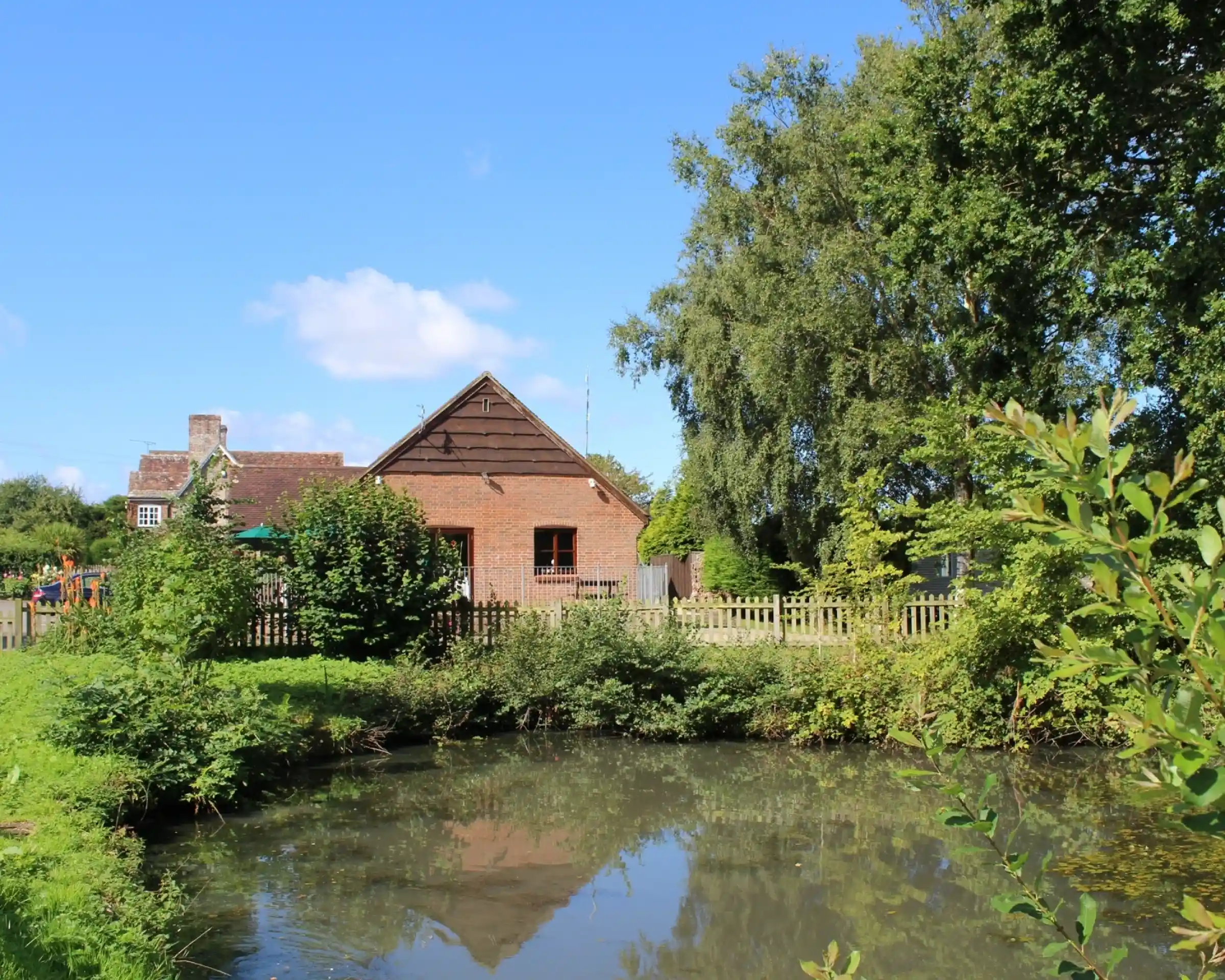 A quaint brick house with a sloping roof sits next to a calm pond surrounded by greenery. The clear blue sky and scattered clouds create a serene atmosphere.