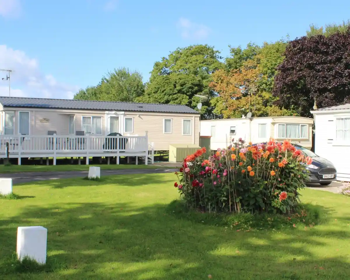 A serene outdoor scene featuring several mobile homes surrounded by lush green grass and a vibrant flower bed with colorful blooms. Trees with autumn foliage are visible in the background against a clear blue sky.
