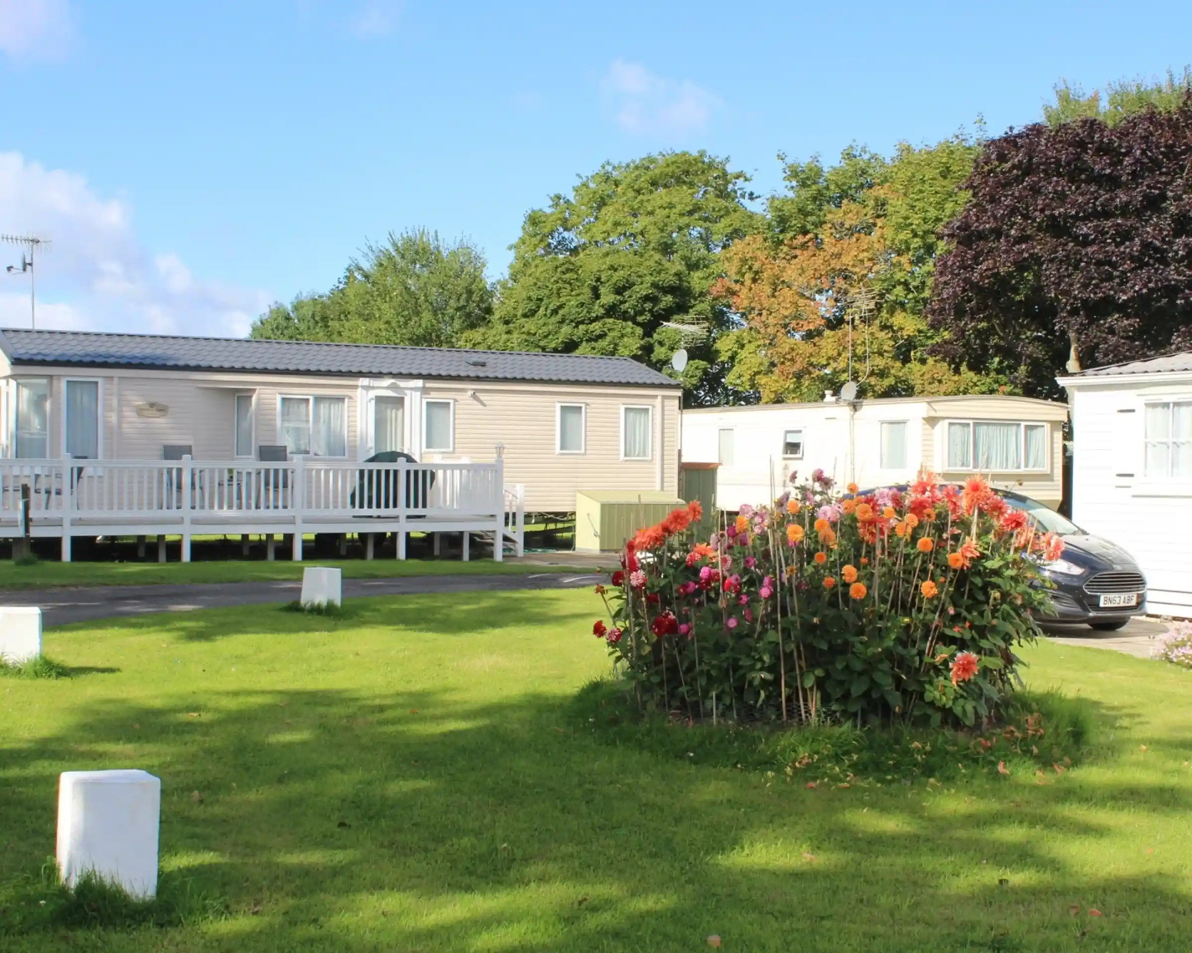 A serene outdoor scene featuring several mobile homes surrounded by lush green grass and a vibrant flower bed with colorful blooms. Trees with autumn foliage are visible in the background against a clear blue sky.