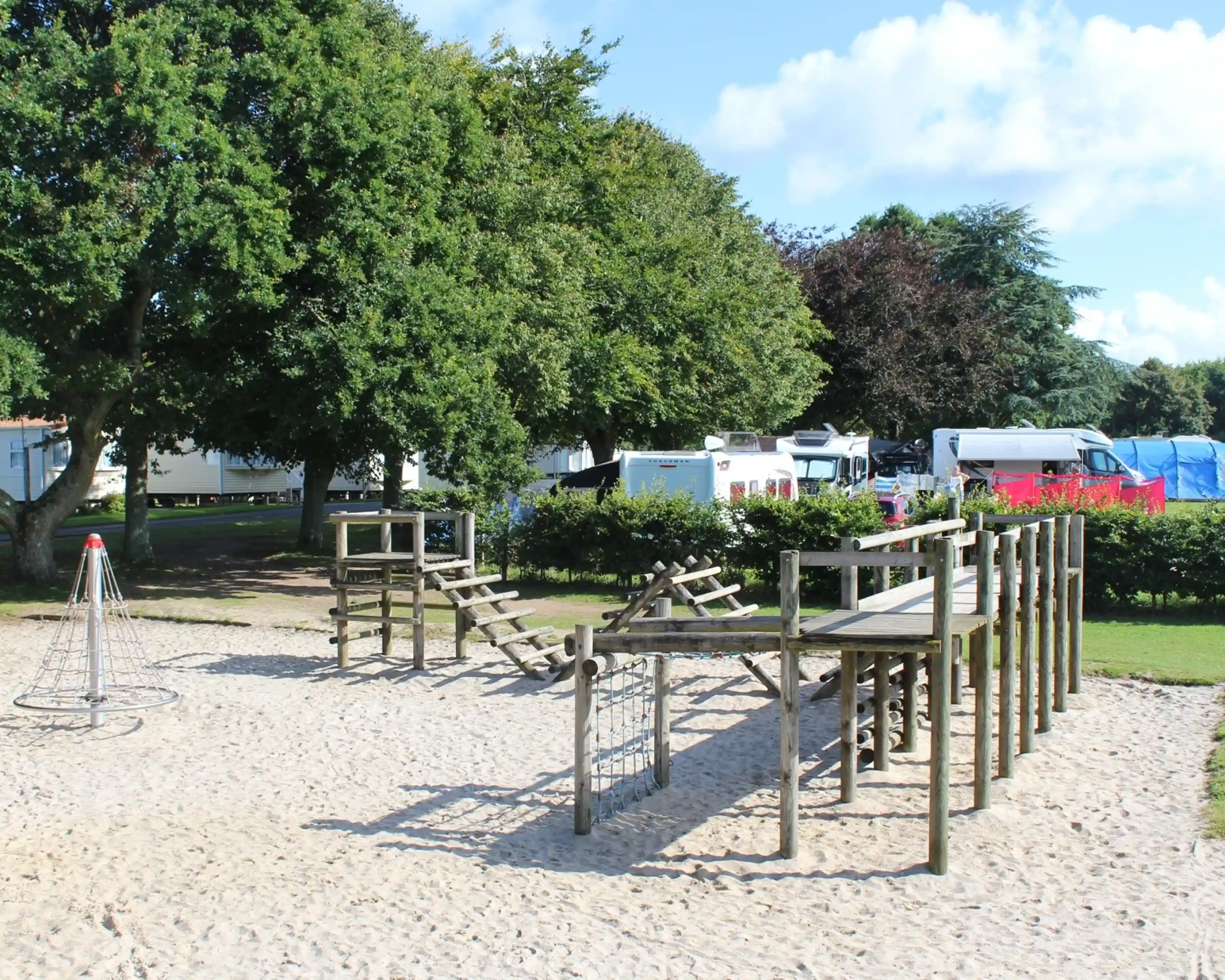 A children's playground featuring wooden climbing structures, a rope climbing frame, and a sandy area, surrounded by trees and camping vehicles in the background.